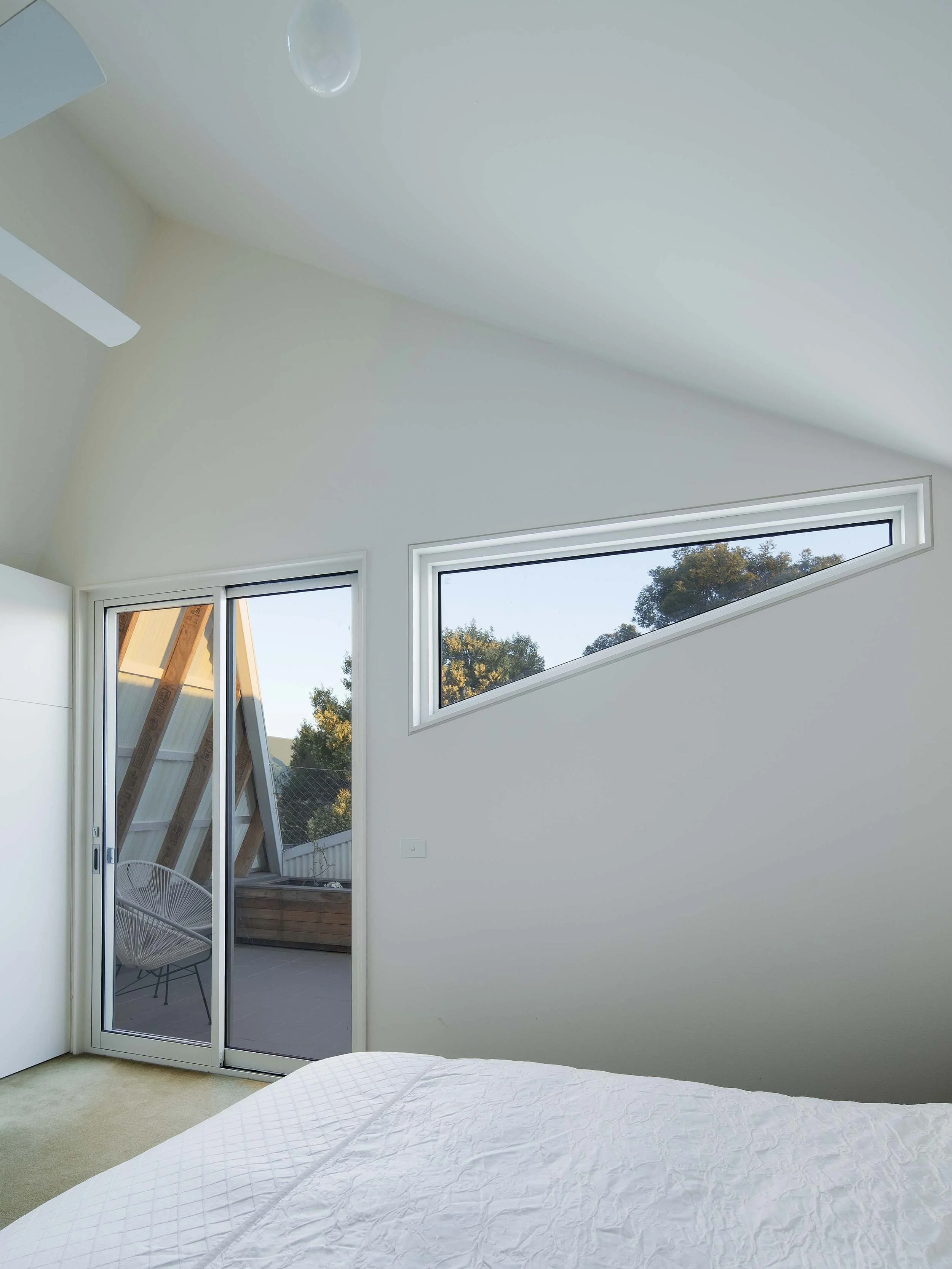 Interior of a modern bedroom with white walls, a bed with a white quilt, and large windows with a view of trees outside. A sliding glass door opens to a small balcony with an outdoor chair and wooden railing. There is a partial view of a sloped ceili
