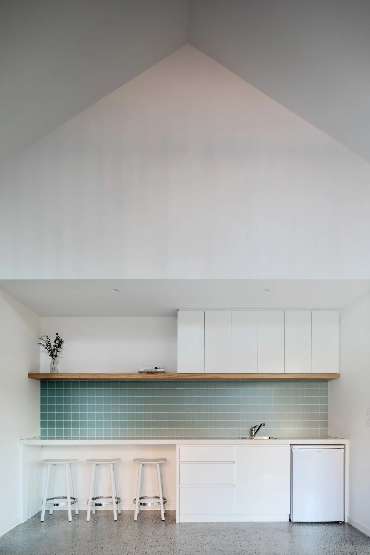 Minimalist kitchen with white cabinets, a light wood floating shelf, turquoise tile backsplash, a small sink, and three white bar stools. There is a small plant and some books on the shelf.