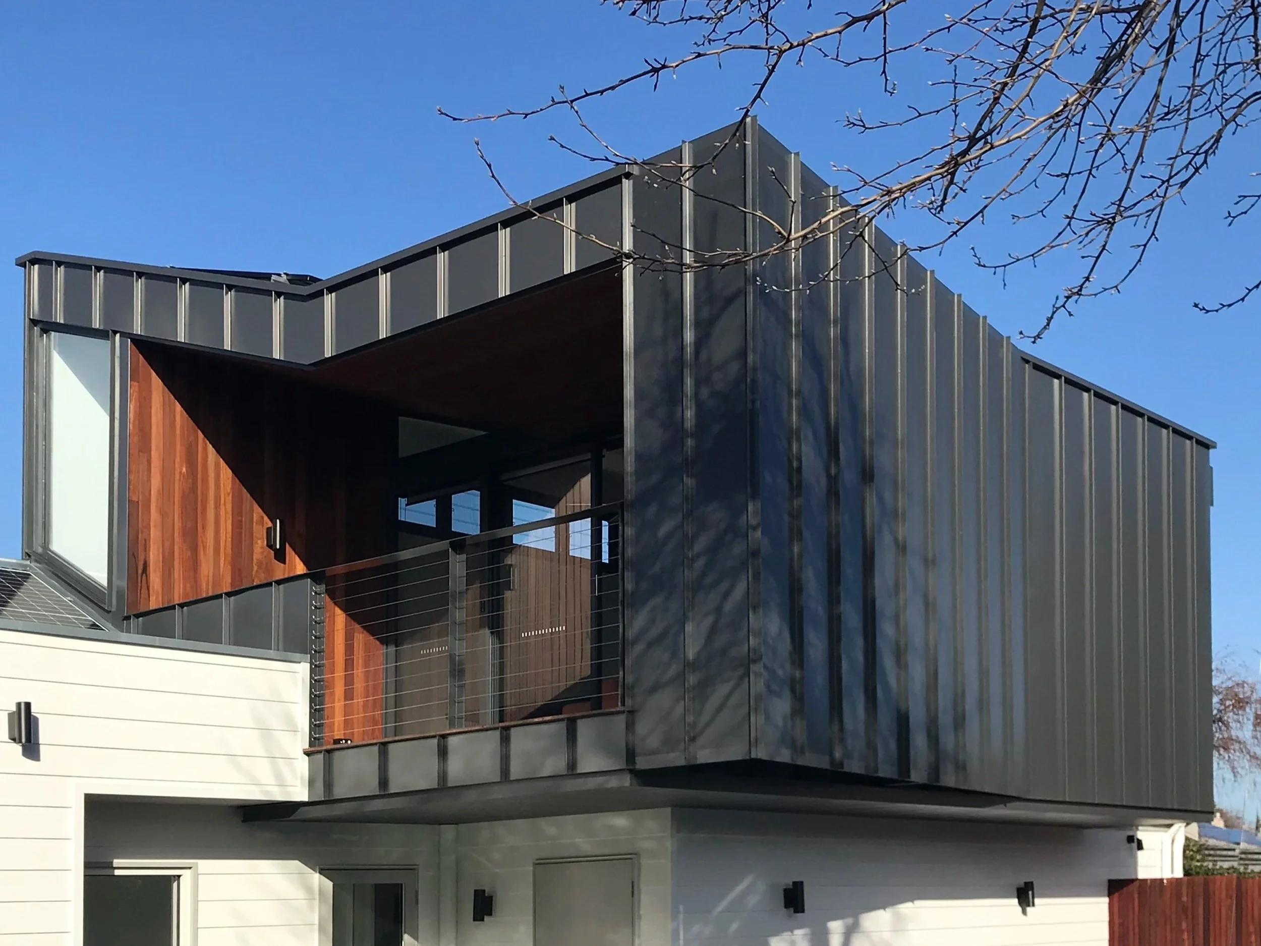 Modern building with metal siding, wooden accents, balconies, and glass doors, against a clear blue sky with tree branches in the foreground.