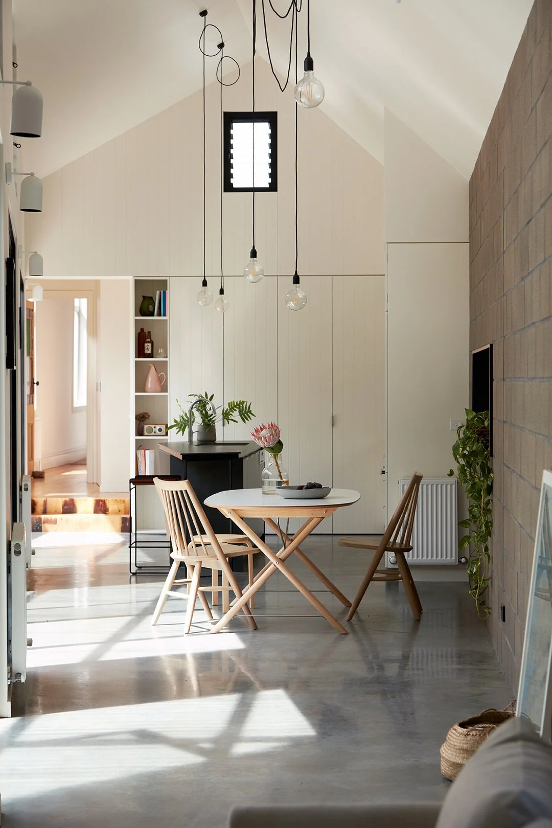 Bright dining area with round white table, three wooden chairs, and hanging light bulb fixtures in a modern, minimalist home.