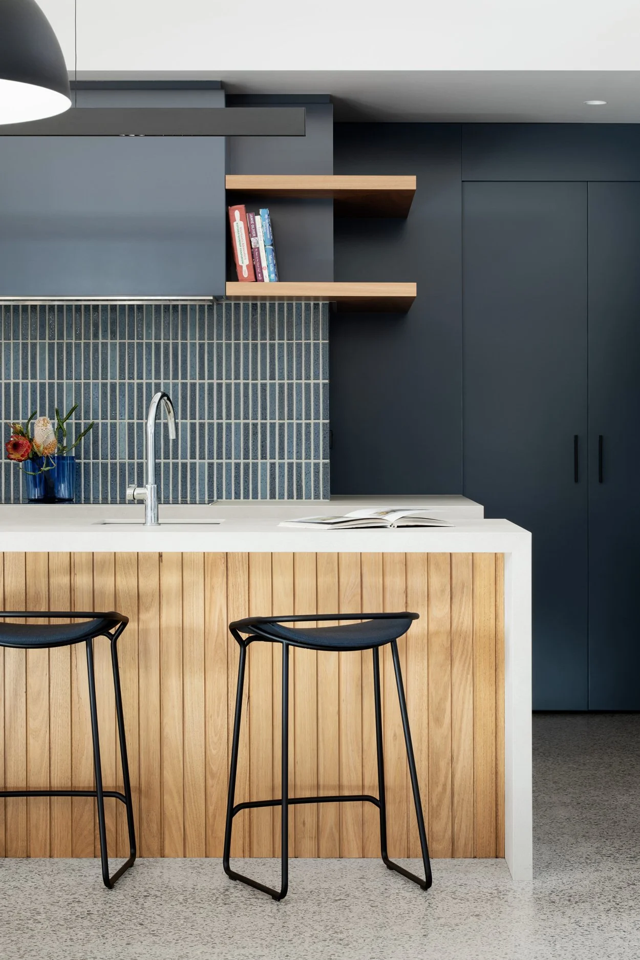 Modern kitchen with a white countertop island, wooden paneling, black stools, blue tiled backsplash, dark blue cabinets, and open shelves with books.