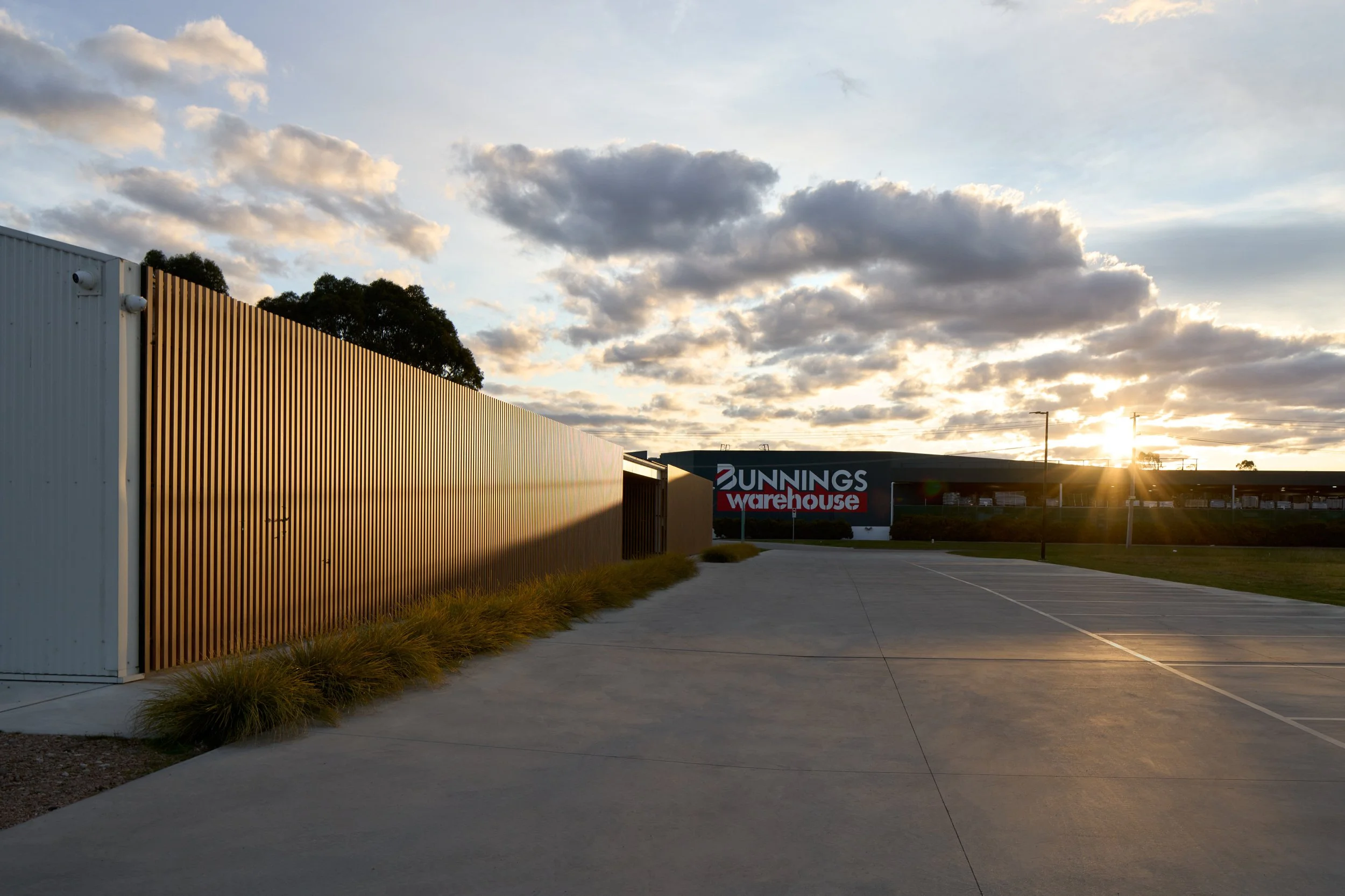Empty parking lot near Bunnings Warehouse with a large metal fence on the left and a partly cloudy sky at sunset.