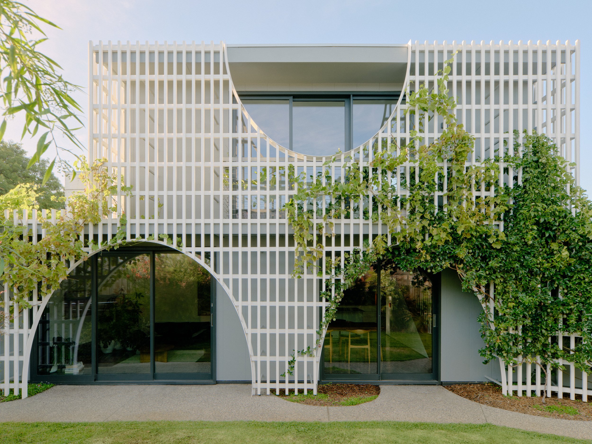 Modern building with white vertical slats, a curved decorative element, and green climbing plants, with large glass doors and concrete walkway in front.