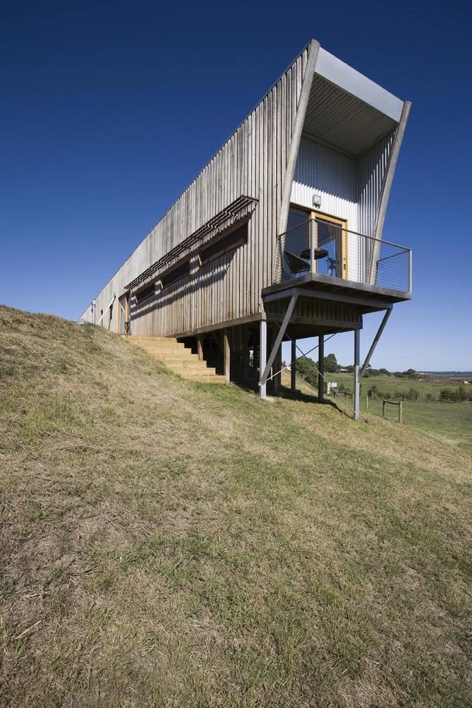 Modern house with wooden exterior and a glass door, built on stilts on a grassy hillside under a clear blue sky.