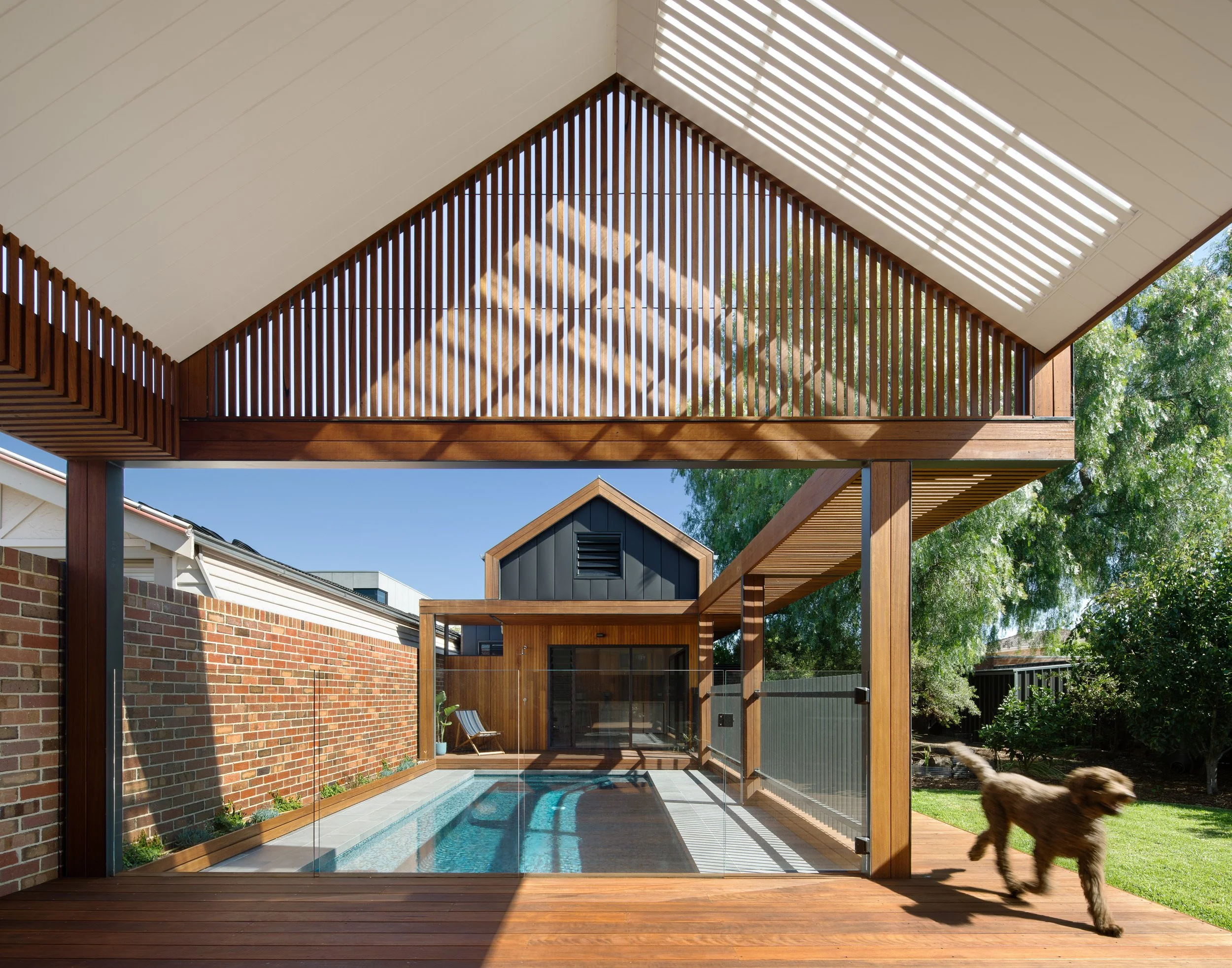 Modern backyard with a small swimming pool, wooden deck, and a wooden and brick outdoor structure with a slatted roof, surrounded by green trees.
