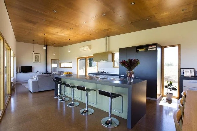 Open-concept kitchen and living room with wooden ceiling, white walls, and large windows, featuring a kitchen island with four black bar stools and a vase of flowers.