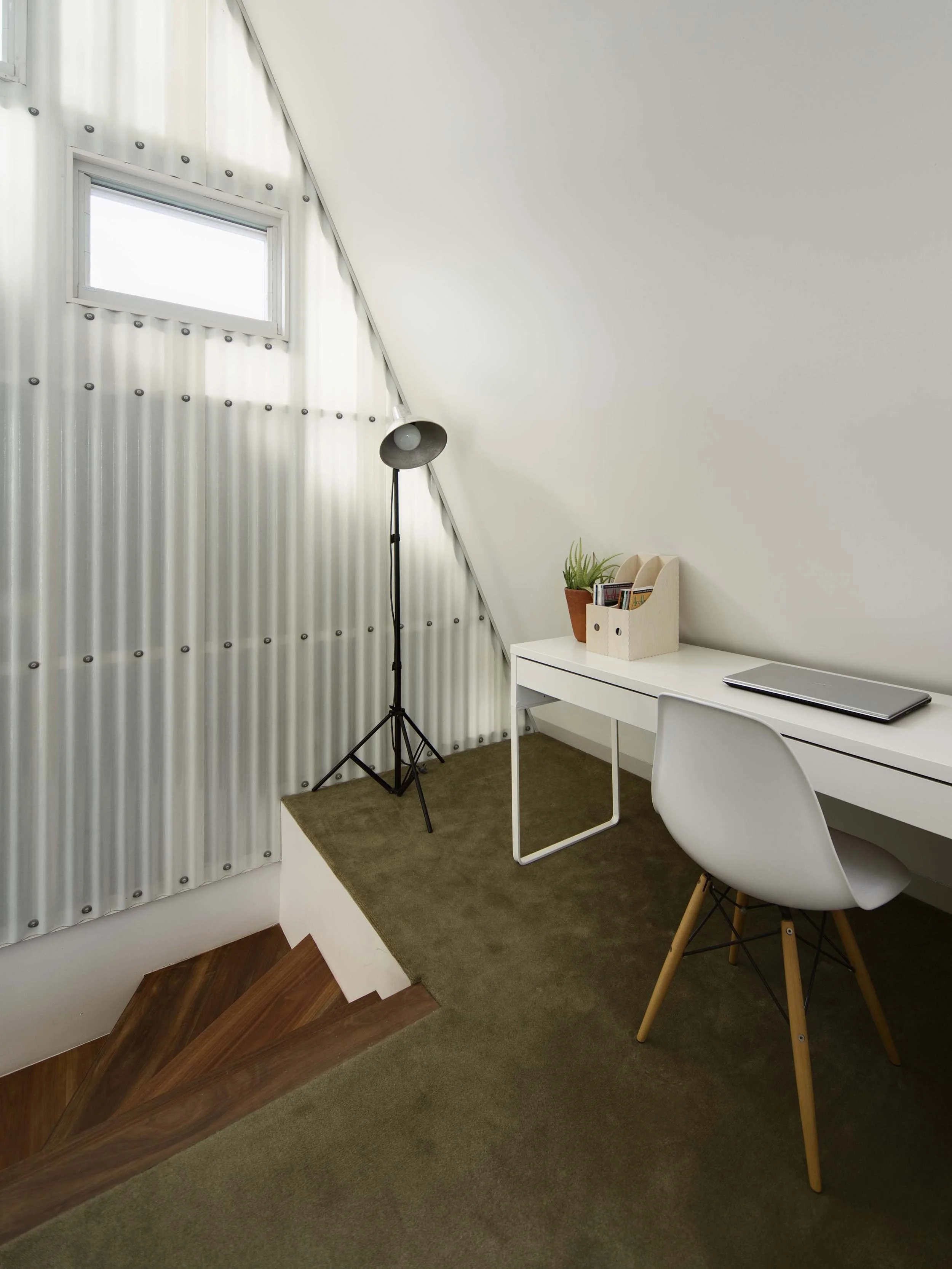 Interior view of a room with a sloped ceiling, small rectangular window, metal wall paneling, a standing floor lamp, a white desk, a white chair, a closed laptop, and a small shelf with books and a potted plant, on beige carpeting.