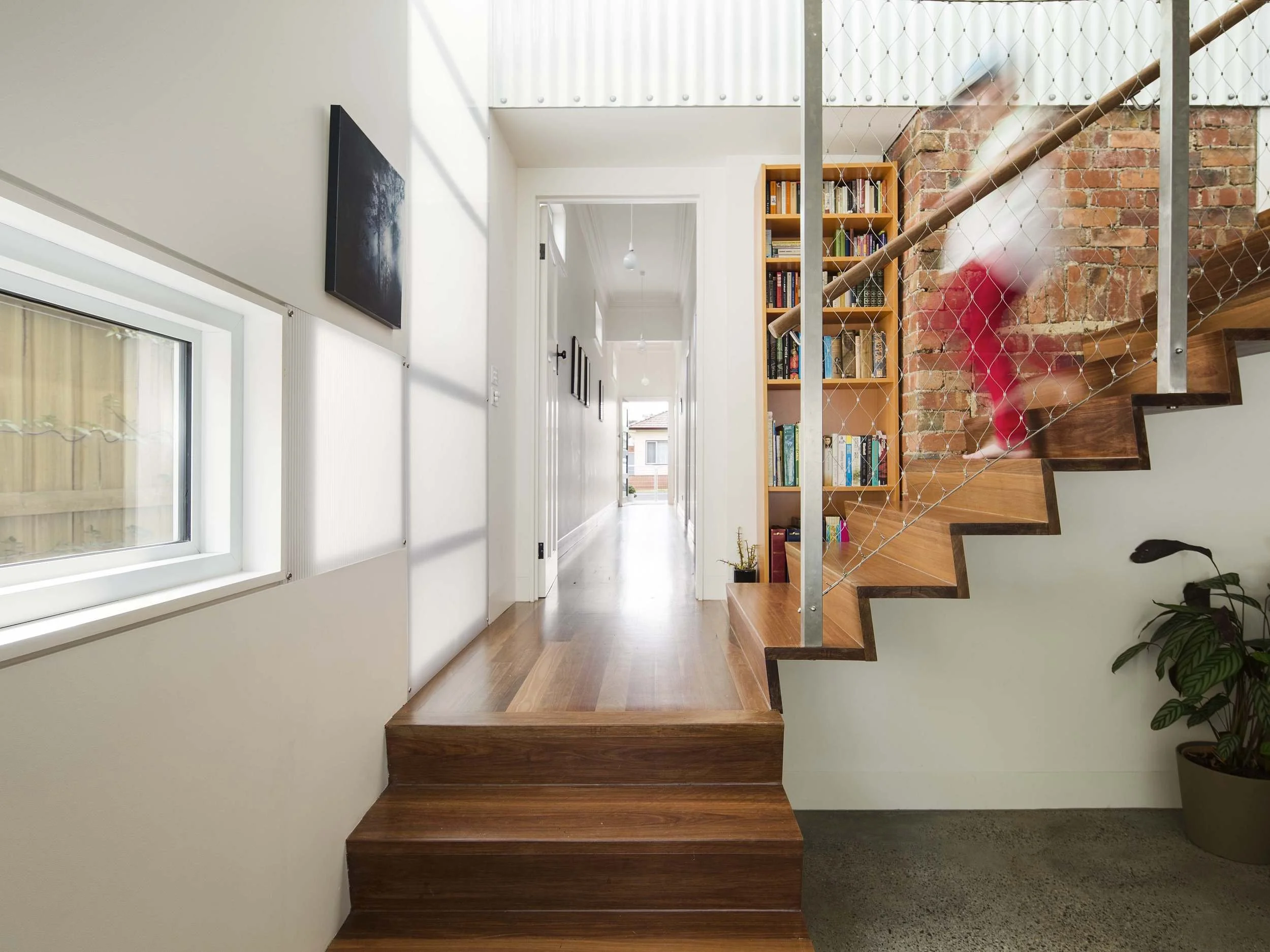 Interior view of a modern home with wooden stairs, a bookshelf, a hallway, and a large window bringing in natural light.