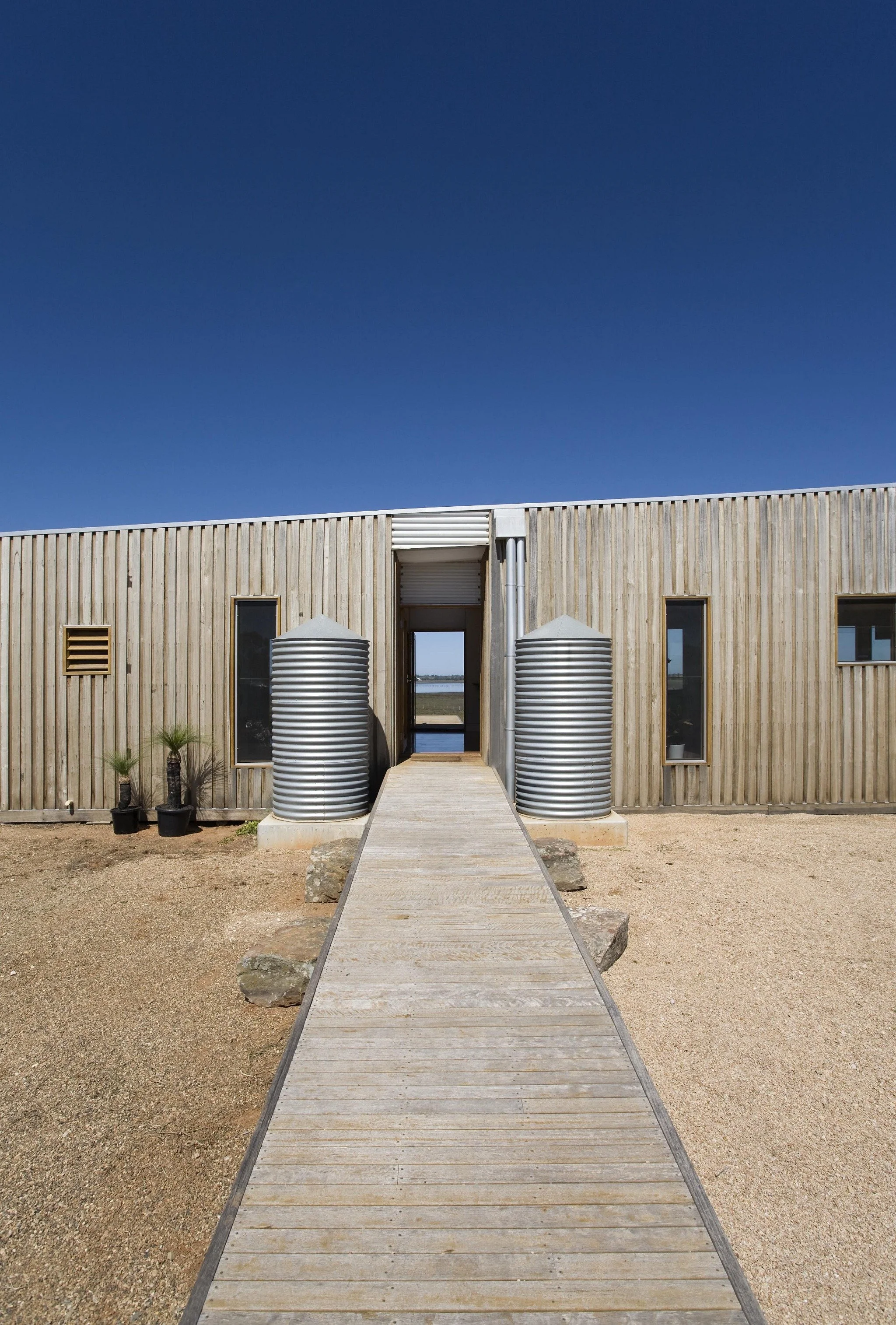 Modern wooden building with a walkway leading to an open doorway, flanked by two large metal water tanks, with small potted plants on the left, under a clear blue sky.