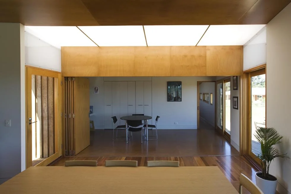Interior view of a modern room with wooden flooring, large windows, a dining table with chairs, and ceiling lights.
