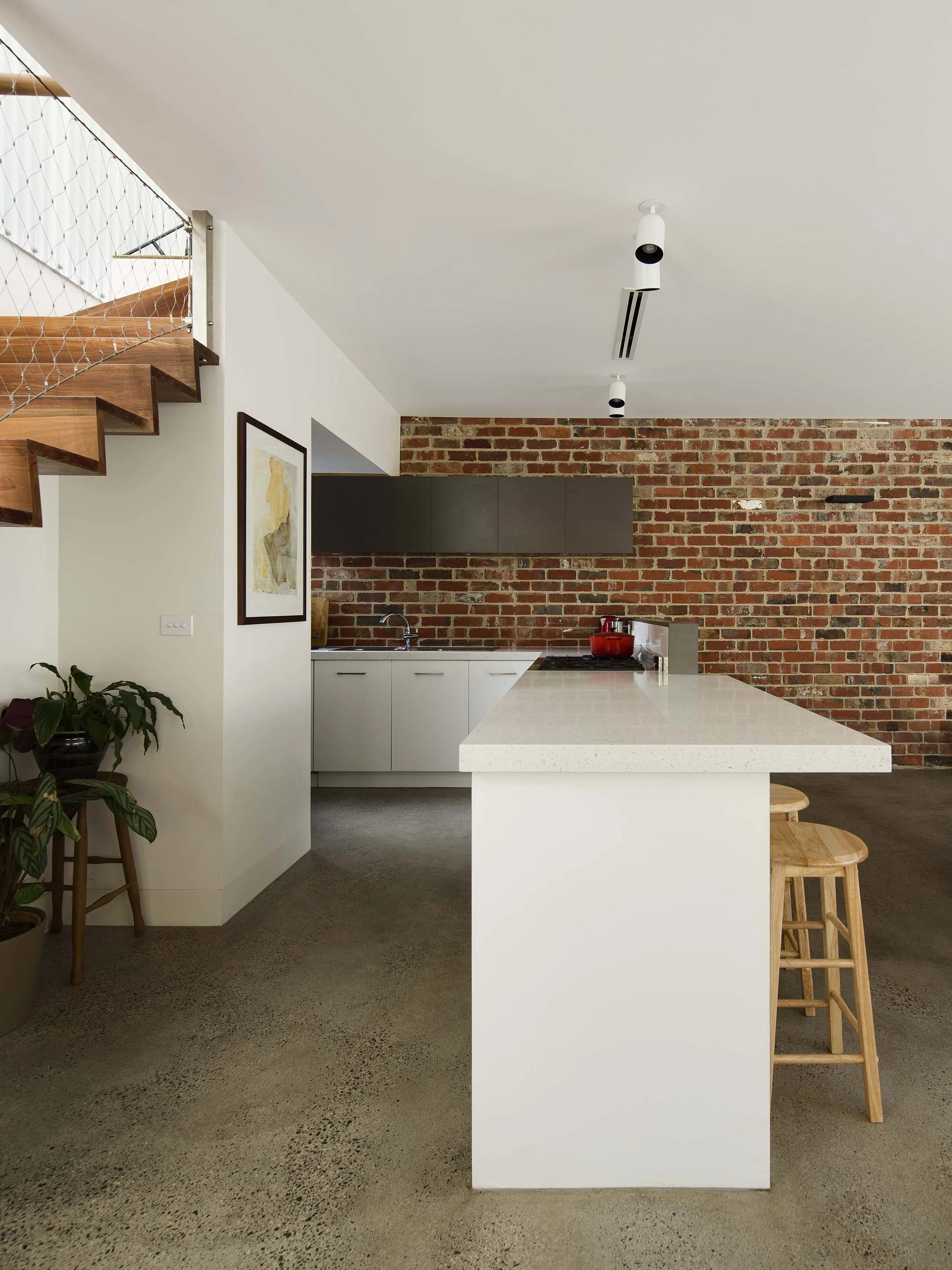 Modern kitchen with white island, brick wall, black cabinets, pendant lights, wooden stools, potted plant, artwork, staircase with wooden steps and metal railing.