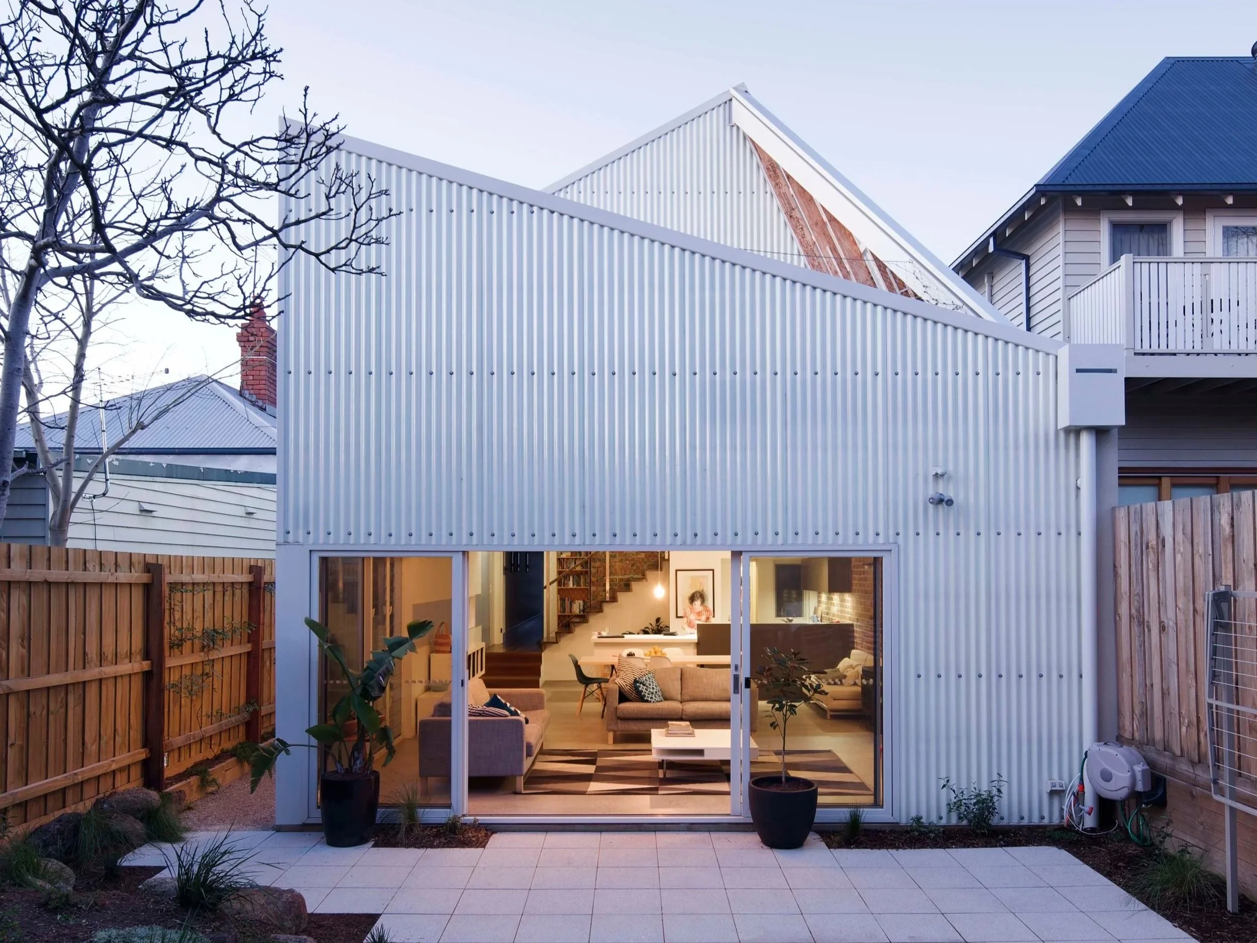 Interior view of a modern home with large glass doors opening to a backyard patio with potted plants. The living room has a beige couch, a coffee table, and wall art, visible through the open doors. The outdoor area features a wooden fence, paving stones, and several plants, with a tree on the left side of the image.