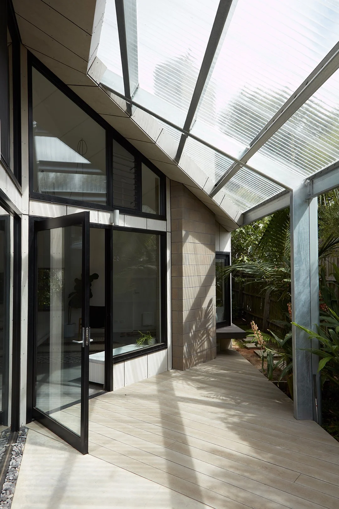 Modern house with large glass windows and a glass roof, surrounded by greenery and a wooden deck.