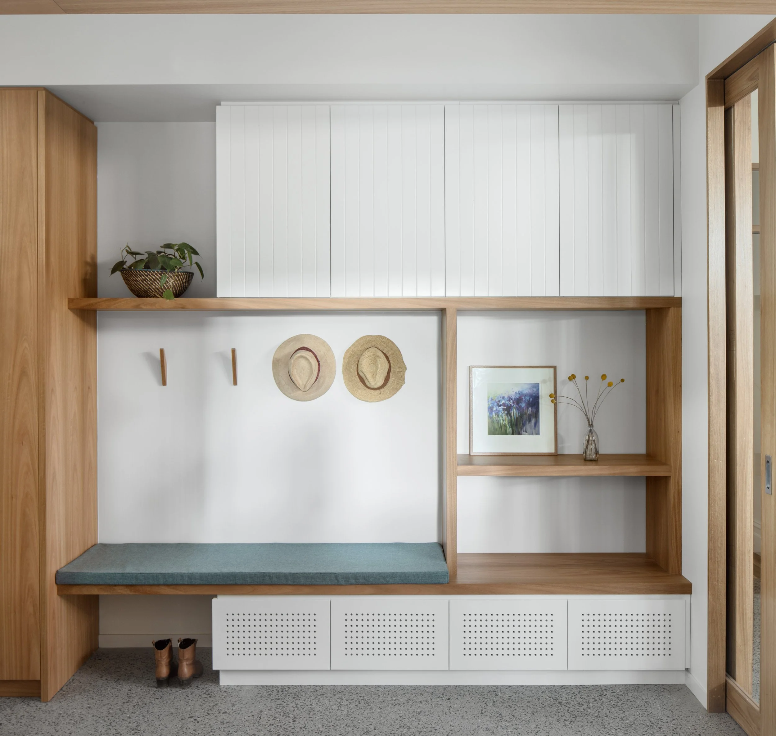 Minimalist interior with a built-in wooden structure, white cabinet, framed art, and hats on a wall hook, with a pair of boots on the floor.