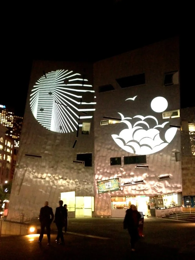 Nighttime view of a modern building with two large light projections on its exterior walls, one resembling a spiral clock and the other showing abstract waves and a bird, with people walking nearby.