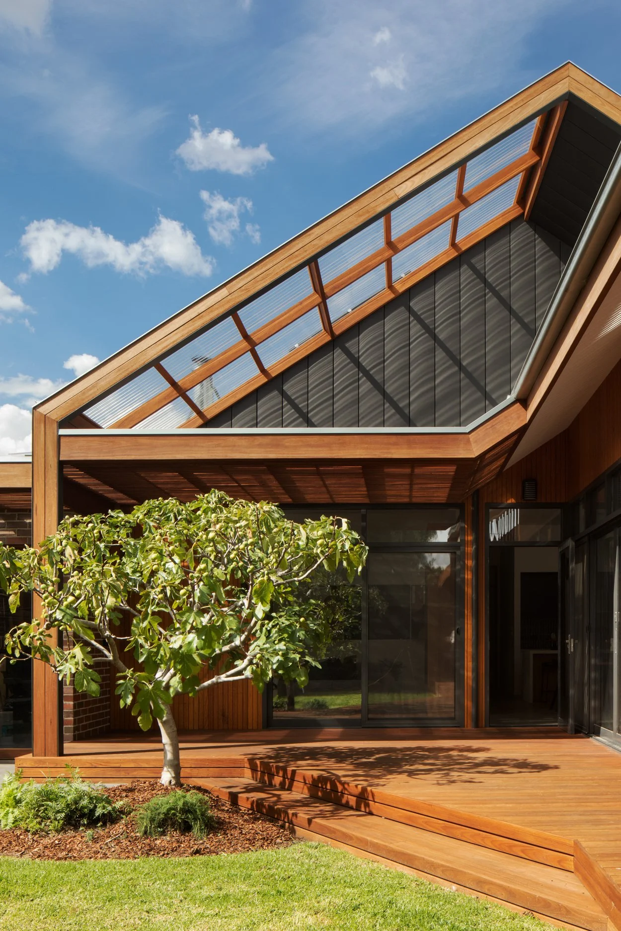 Modern house with large glass sliding doors, a wooden deck, a tree, green grass, and a partly cloudy sky.
