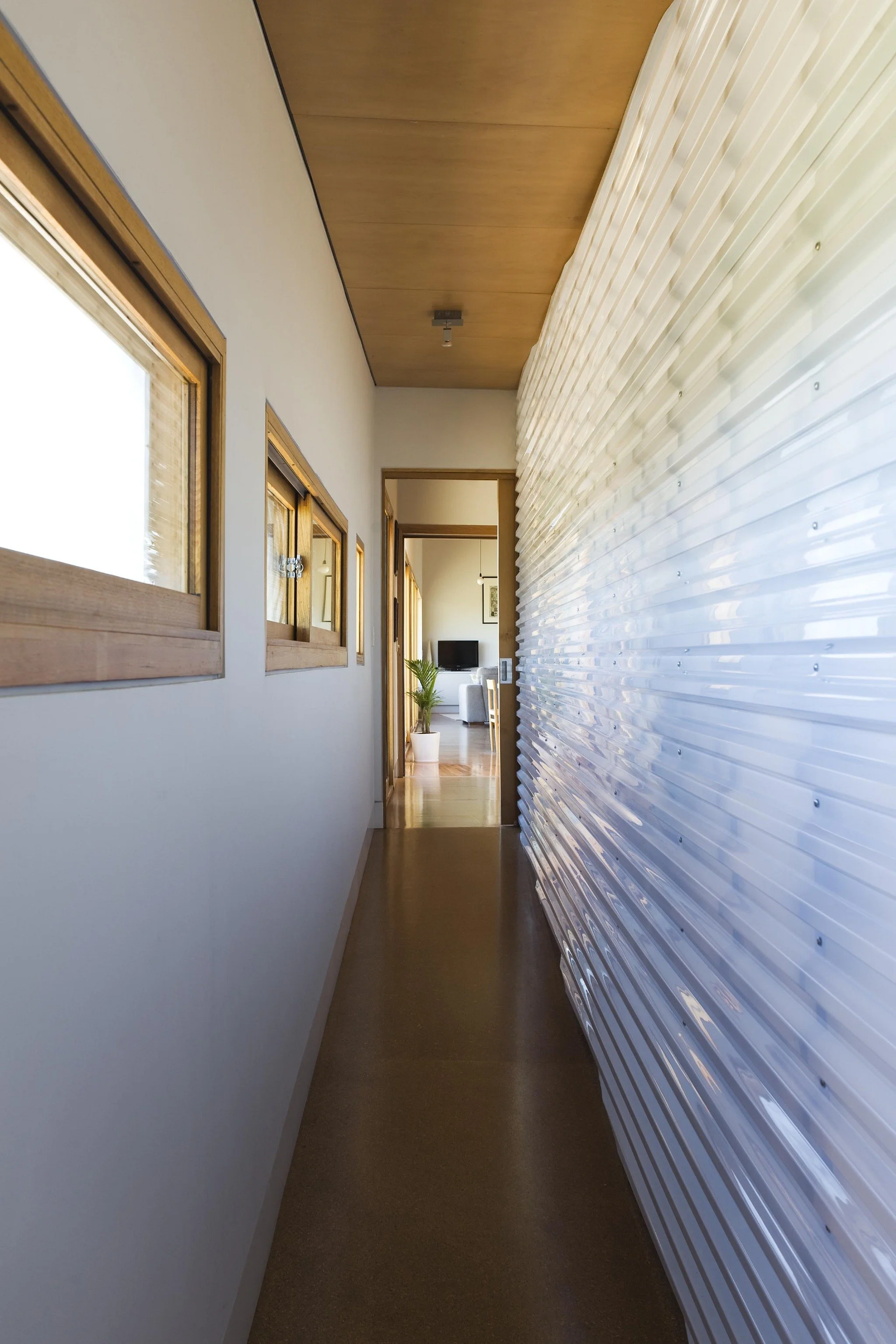A narrow hallway with white walls, wooden window frames, a wood-paneled ceiling, and a large, textured, semi-transparent wall on the right side. At the end of the hallway, there is a small room with a television, a potted plant, and some furniture.