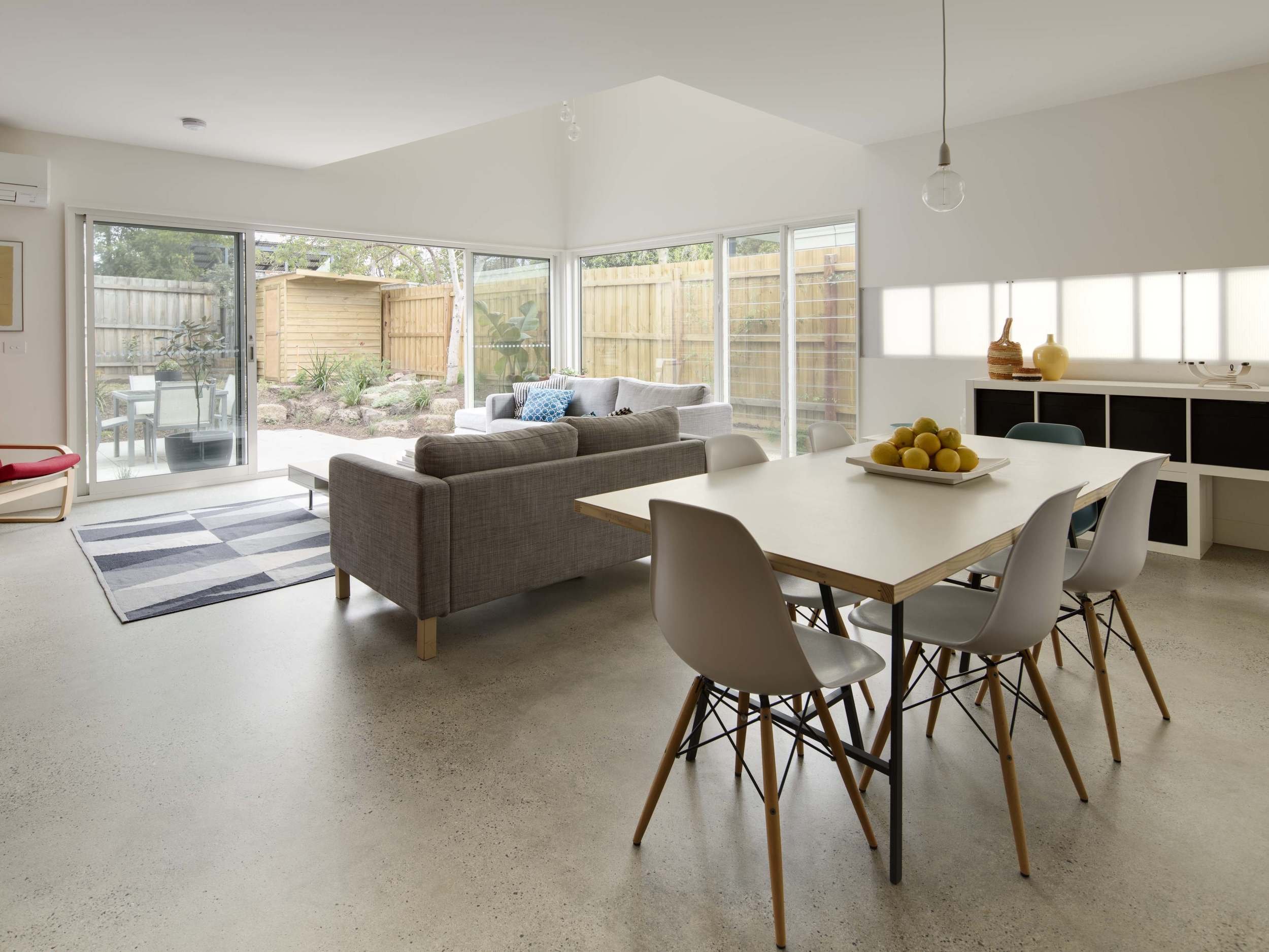 Interior of a modern living room with large windows, a gray sofa with throw pillows, a white dining table with white chairs, a black cabinet with decorative vases, and a view of a backyard garden.