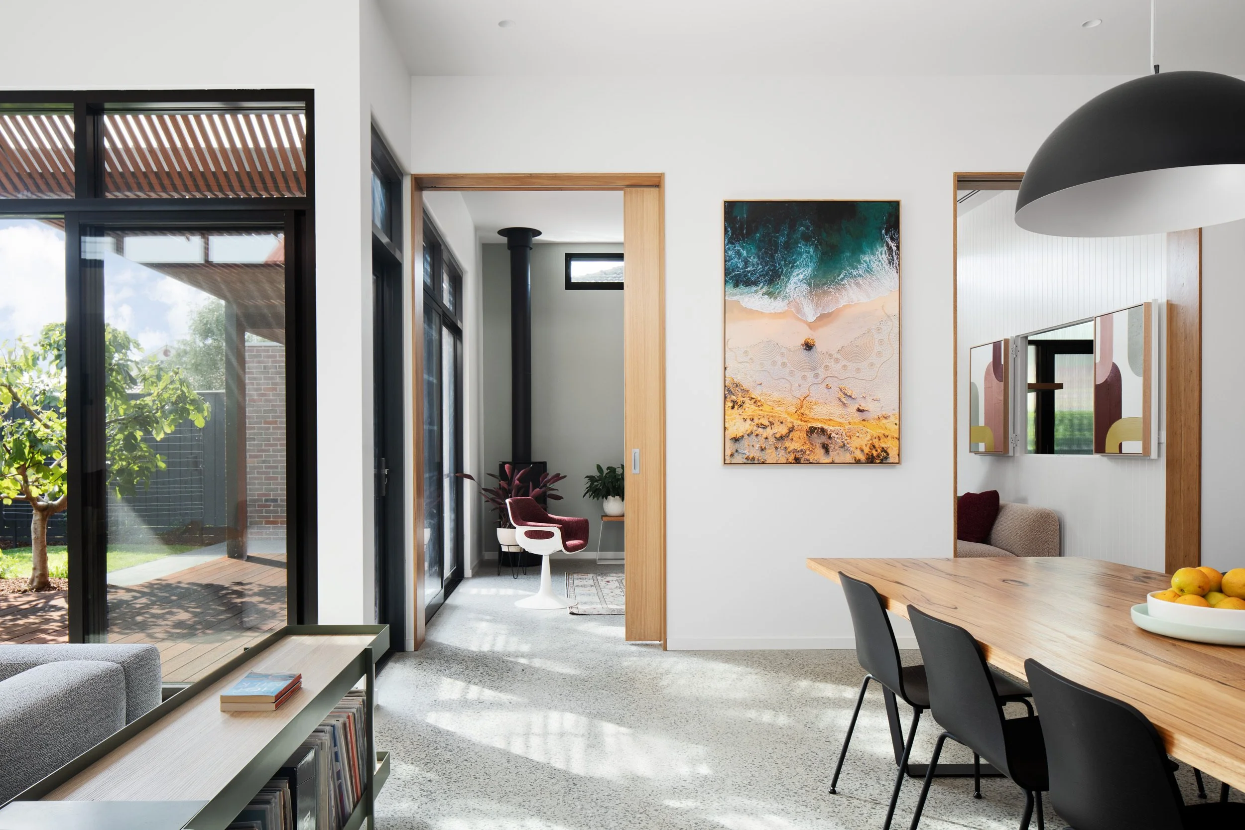 Modern interior living space with large glass doors showing a backyard with greenery, a small room with a white chair and desk, a colorful beach-themed wall art, and a wooden dining table with chairs.