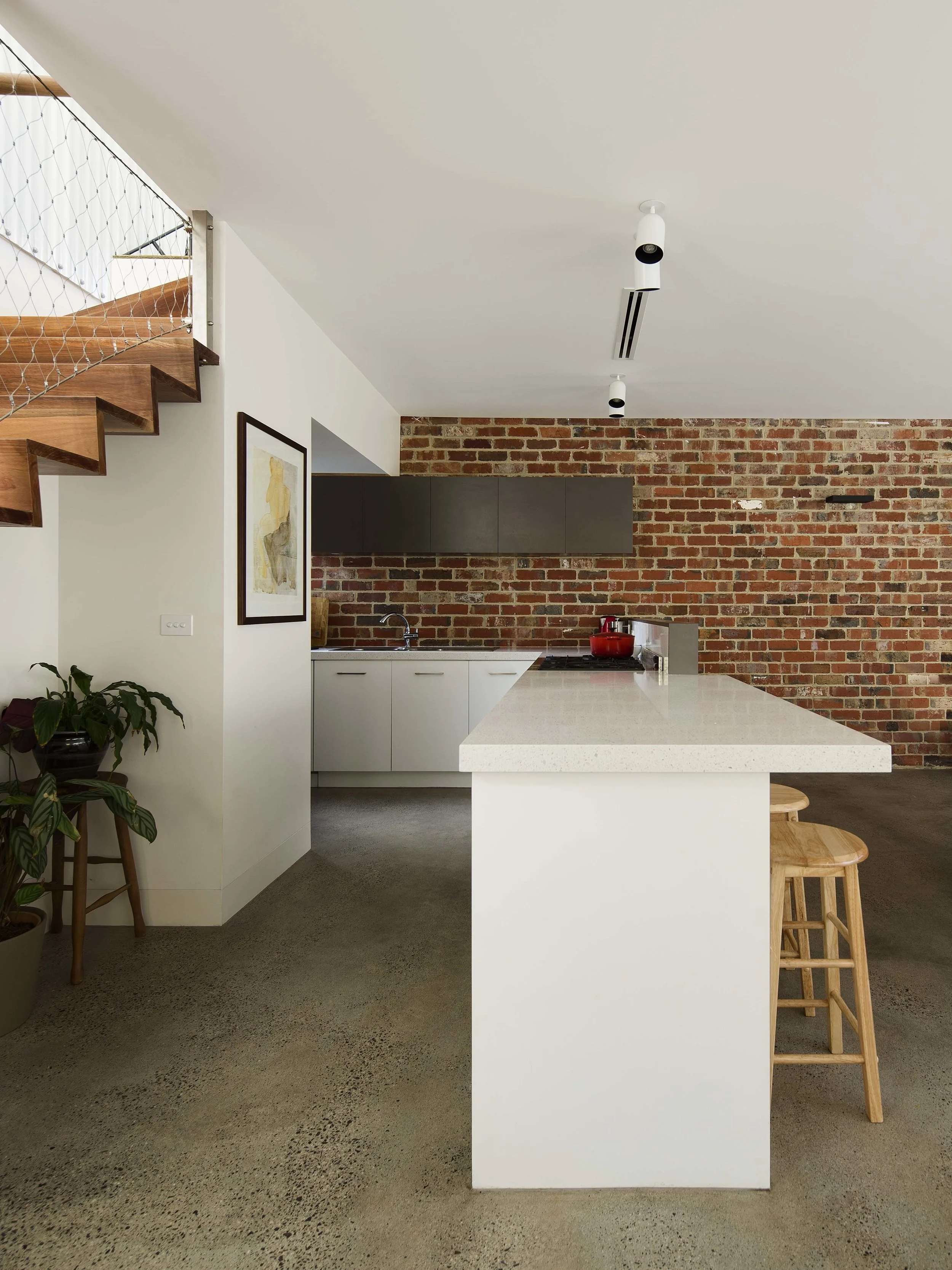 Modern kitchen with a white island, brick wall background, wooden bar stools, and black cabinetry. Includes a plant and staircase nearby.