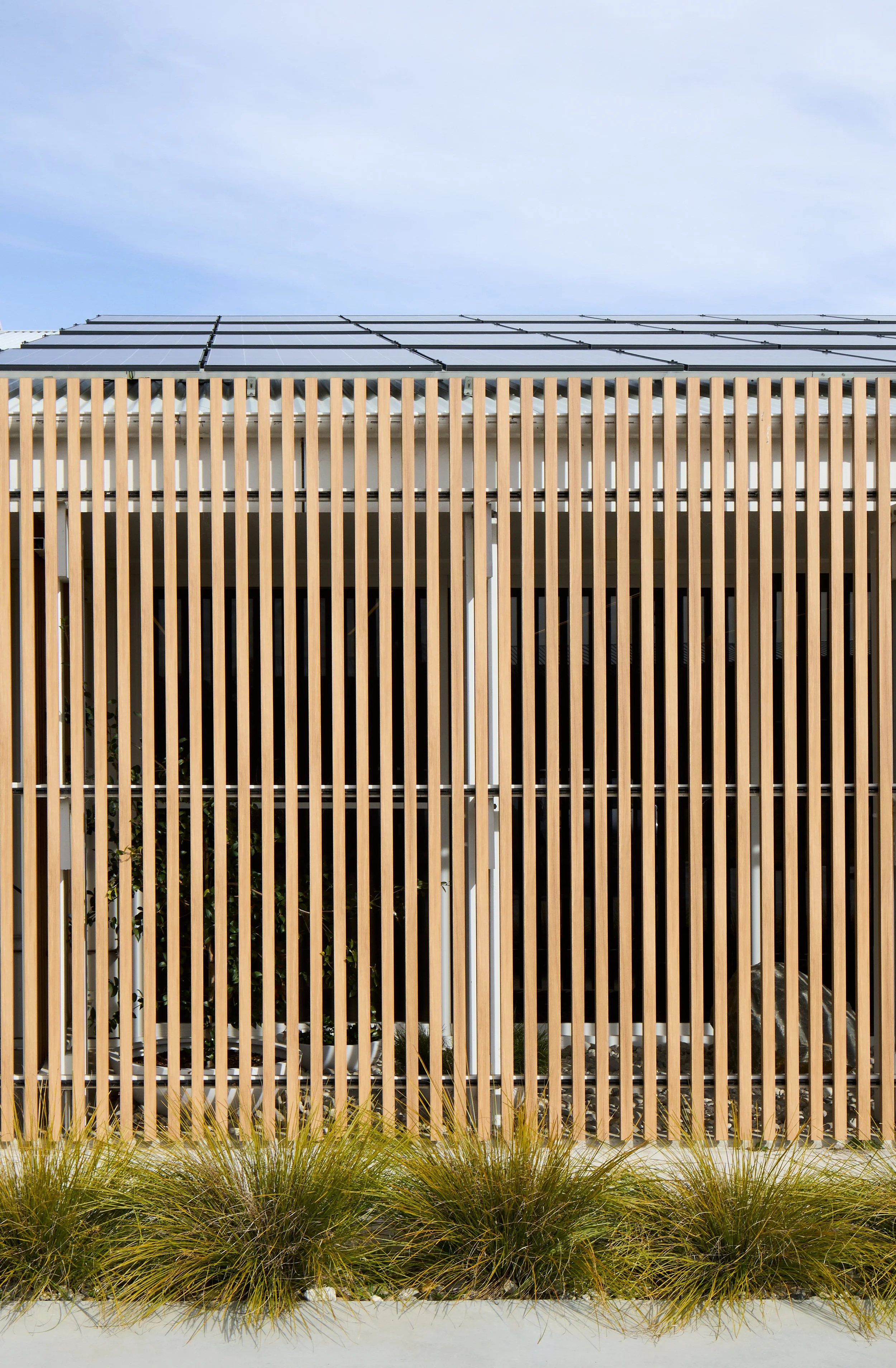 Modern building with a wooden vertical slat fence in front, solar panels on the roof, and tall grass in the foreground.