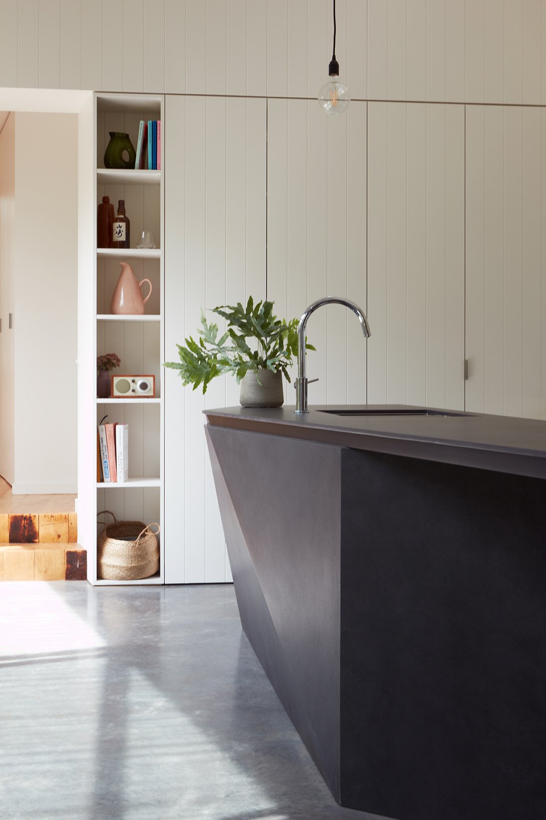 Modern kitchen with a dark gray island, a silver faucet, a plant in a gray pot, white shiplap walls, open shelving with decorative items, wood flooring, and a hanging light bulb
