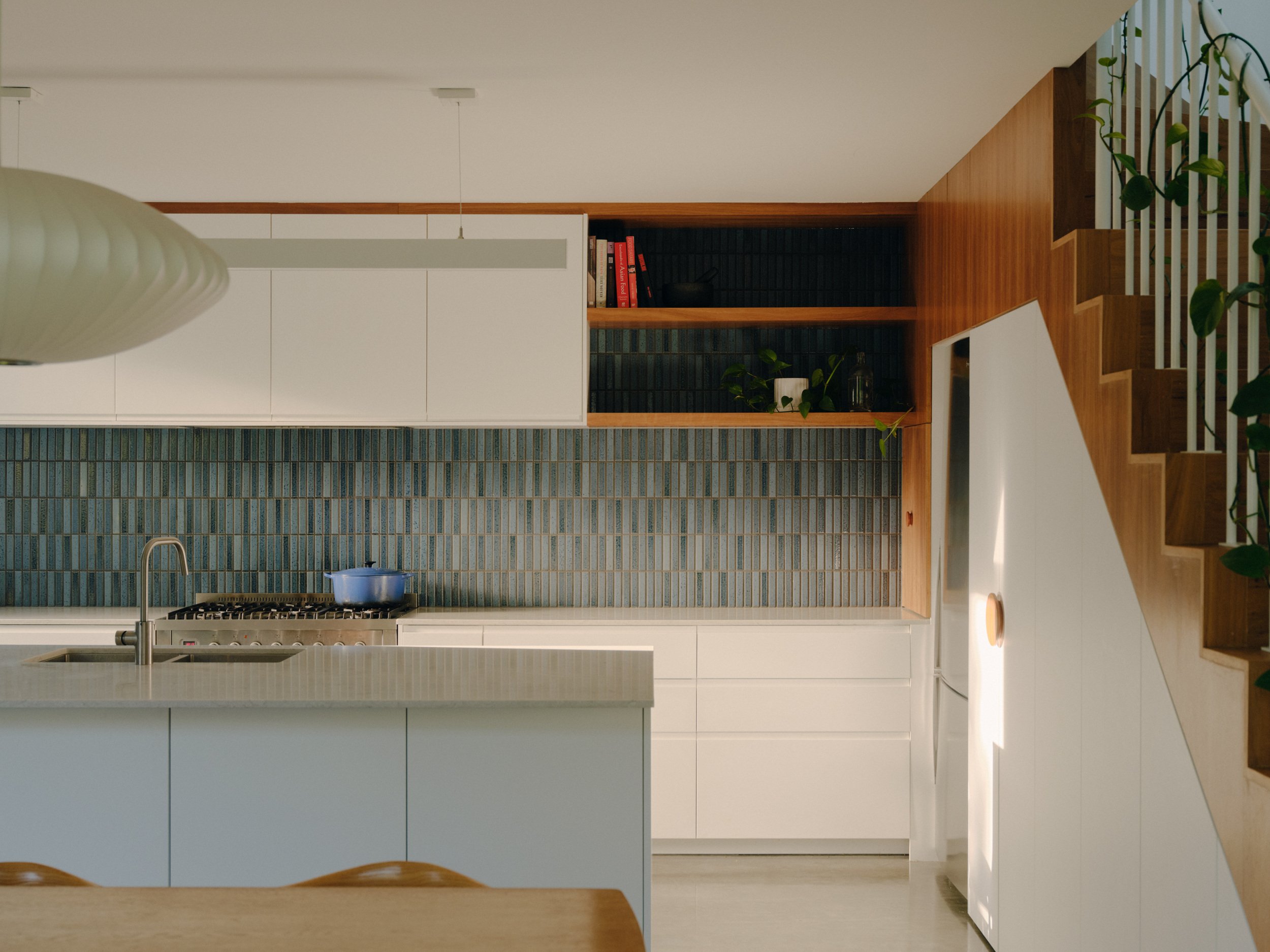 Modern kitchen with white cabinets, a blue pot on stove, hanging vent, and a wooden staircase with green plants.