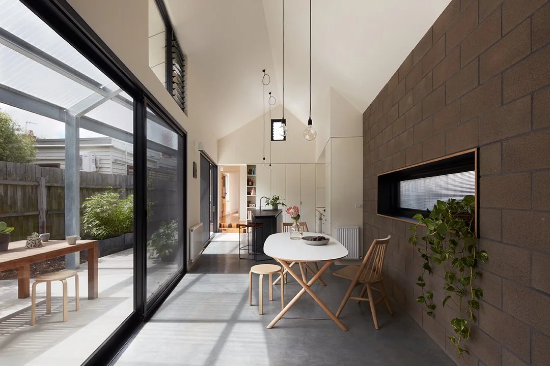 Modern dining area with white table and wooden chairs, adjacent to a large sliding glass door leading to an outdoor patio with wood table and seating, and featuring a brick accent wall with a built-in window and a hanging plant.