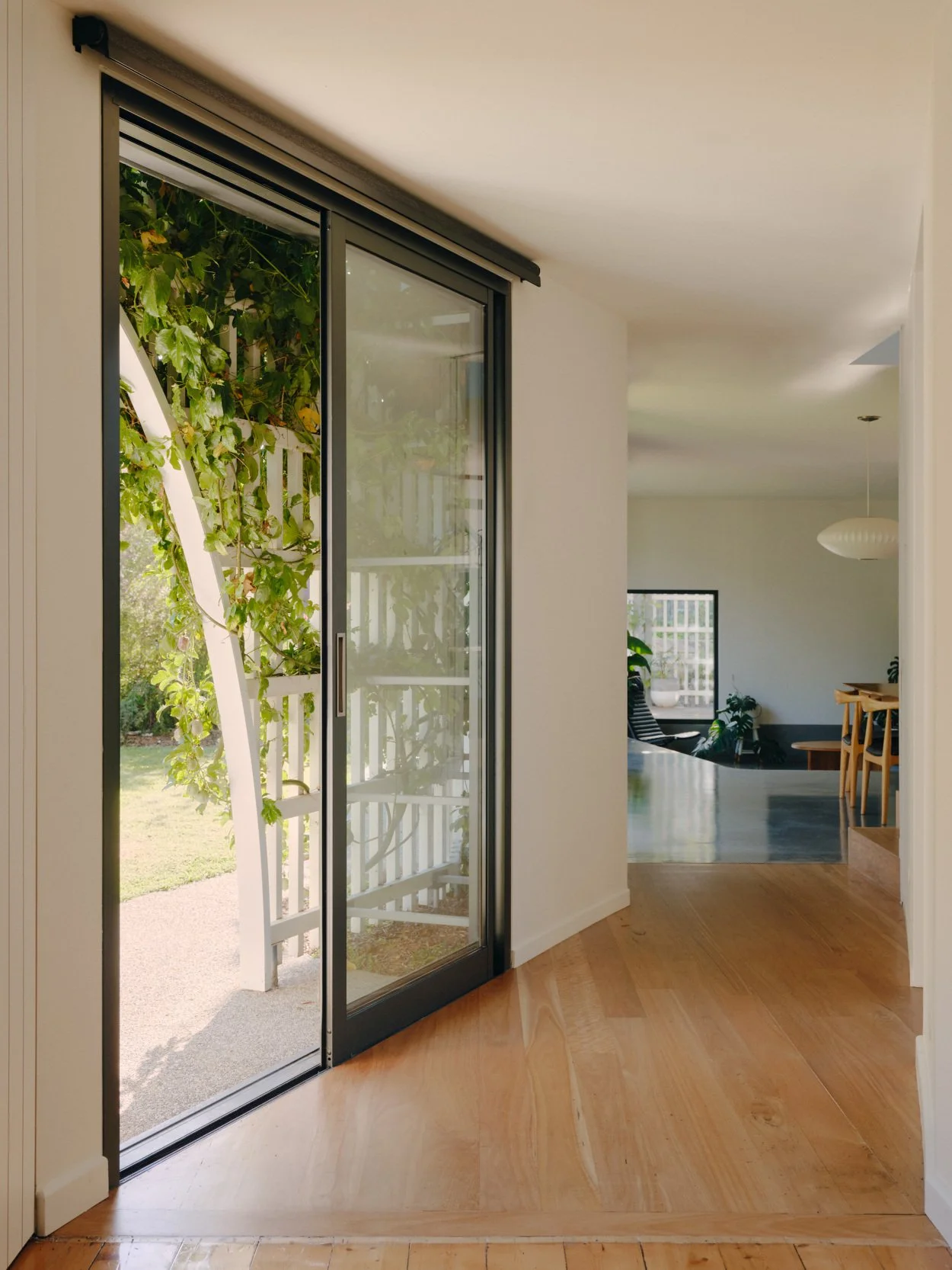 Interior view of a house showing a sliding glass door leading to a garden with green foliage and a white trellis. Inside, there is a wooden floor, a dining area with chairs, a plant, and a window with a grid pattern. The space is brightly lit with na