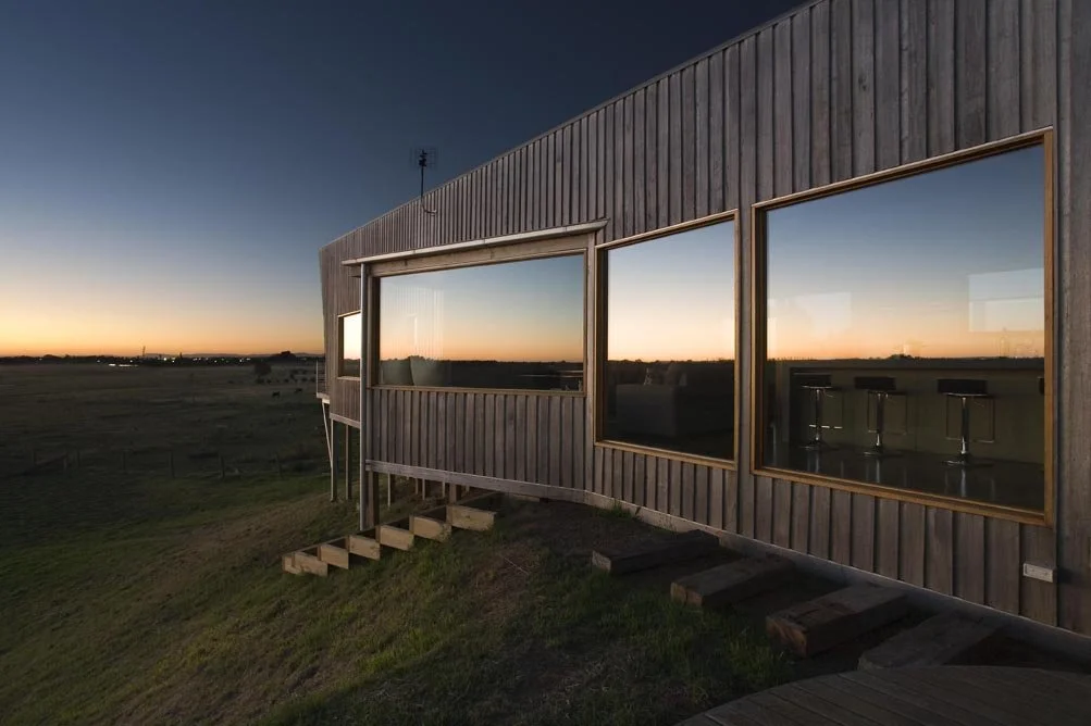 A modern wooden building with large windows overlooking a sunset landscape. The interior of a bar or café with high stools is visible through the windows.