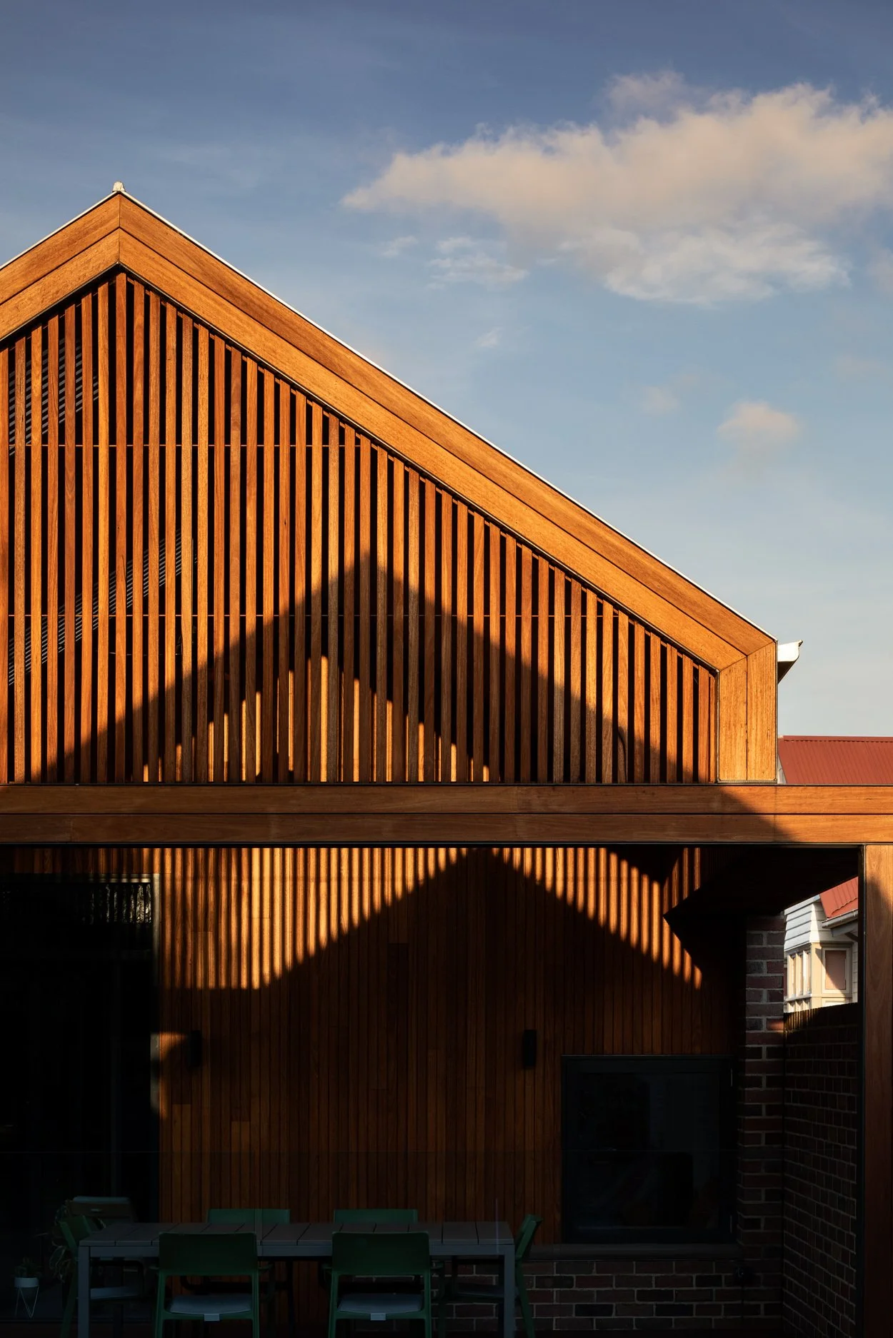 A modern house with a sloped wooden exterior, with shadows cast by the sunlight, and a patio with a table and chairs in the foreground.