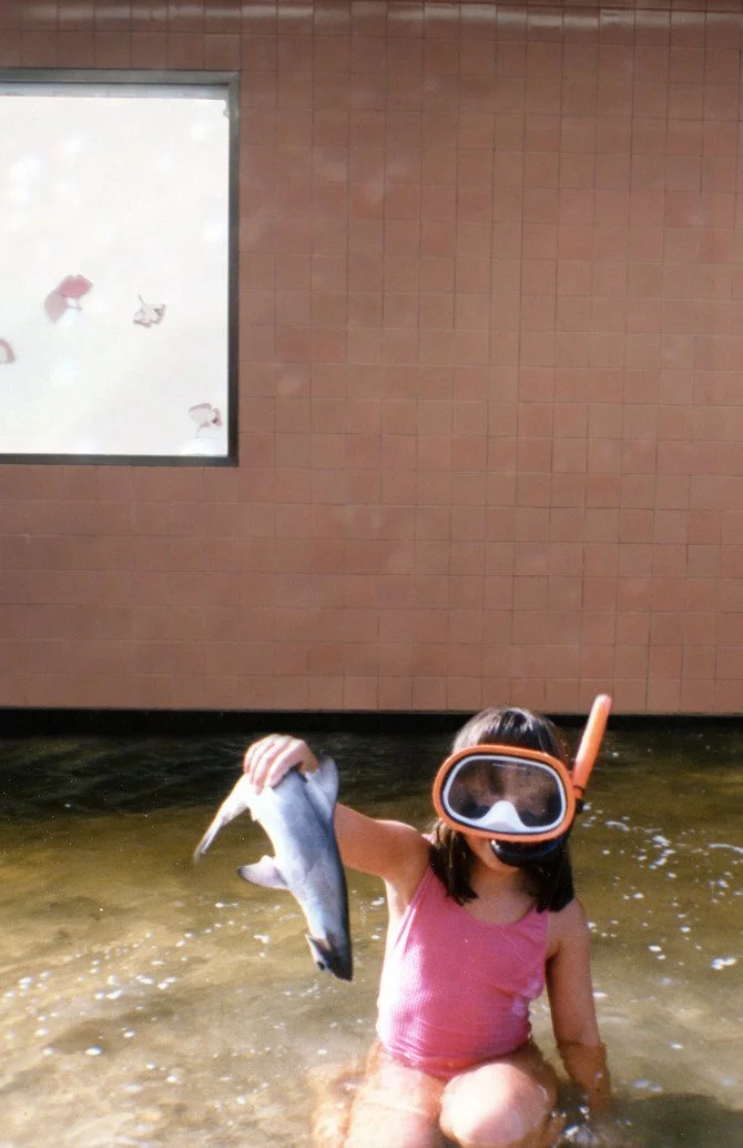 A young girl in a pink swimsuit is sitting in shallow water, holding a small fish in her right hand. She is wearing a snorkel mask and a snorkel tube, and there is a building wall and window behind her.