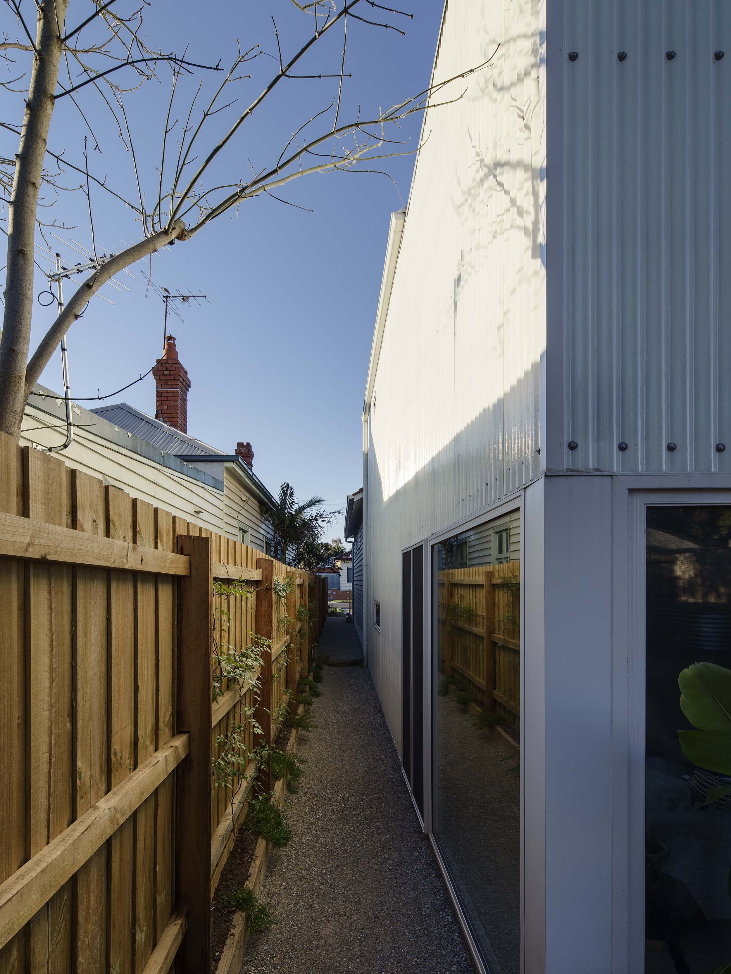 A narrow outdoor alleyway between a wooden fence on the left and a modern building with white and gray paneling on the right. The alley is lined with small plants at the fence base, and a blue sky with a tree and brick chimney visible in the backgrou