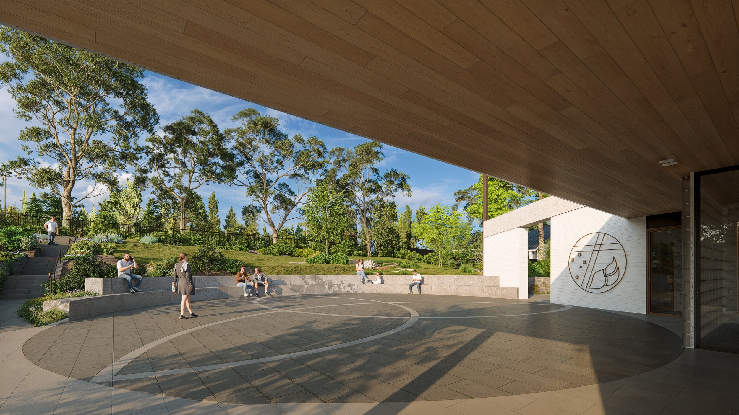 People sitting and walking in an outdoor amphitheater with a grassy area and tall trees in the background, under a large wooden overhang.