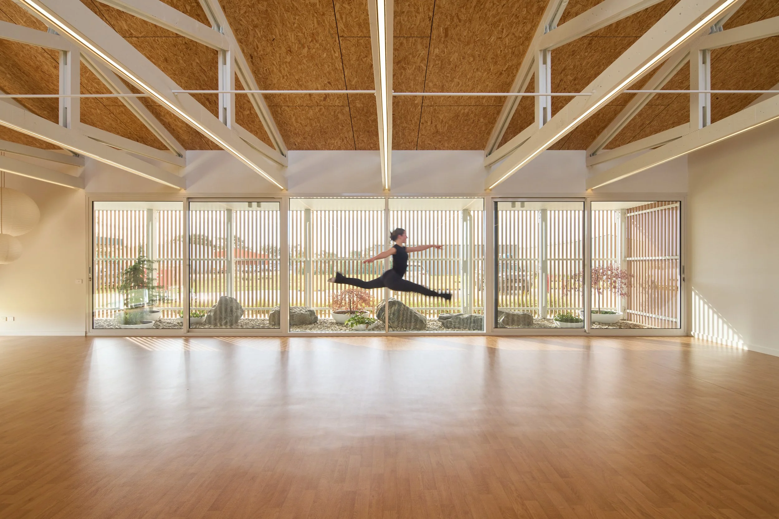 A person practicing ballet or dance in a spacious, modern dance studio with large windows, wooden ceiling, and a light-colored wood floor.