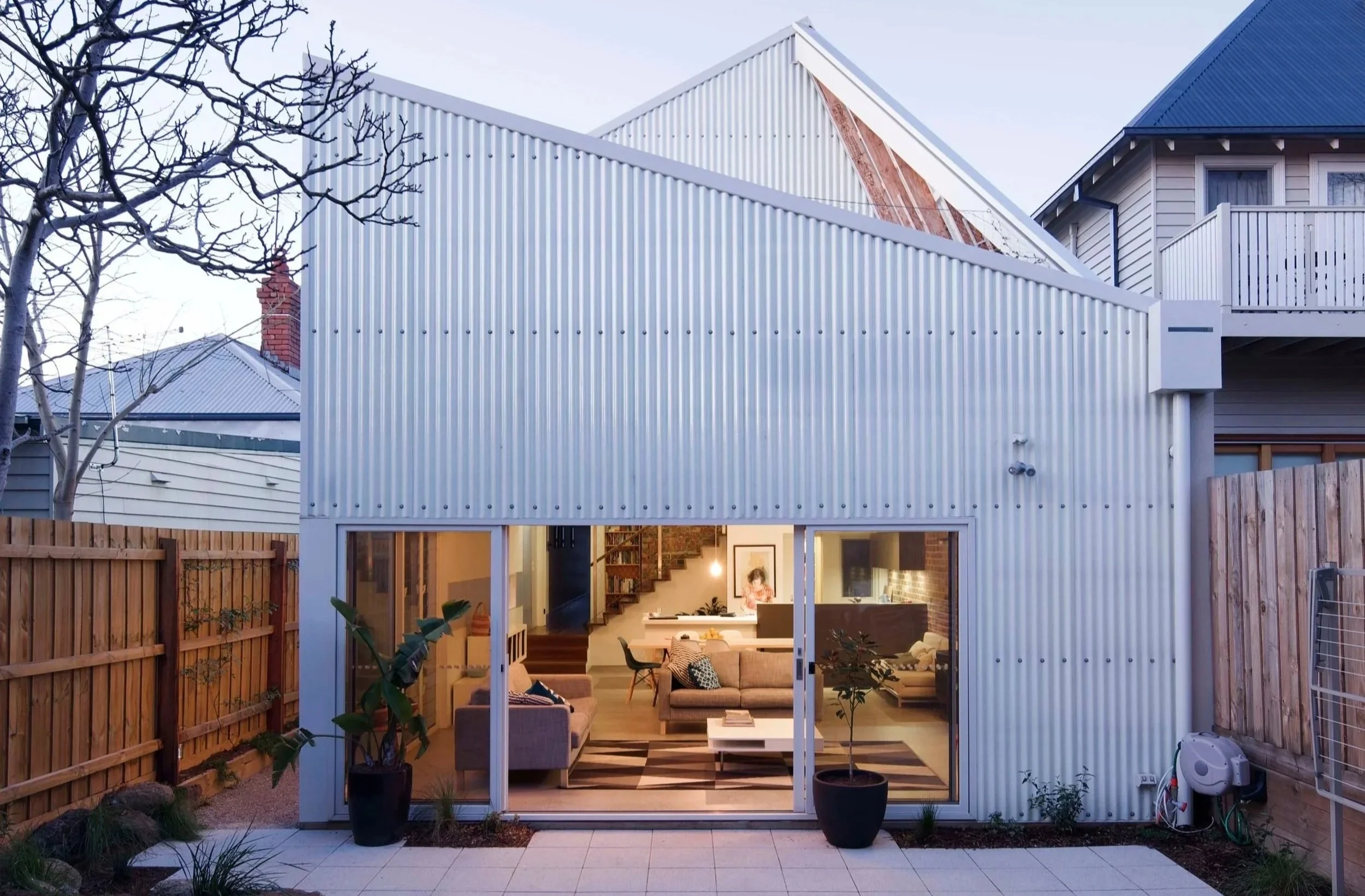 A contemporary house with a metal exterior wall and large glass doors opening to a backyard patio. The interior living space is visible through the doors, decorated with furniture and interior lighting. The backyard has wooden fencing, potted plants, and a tree with bare branches, suggesting it is winter or early spring.