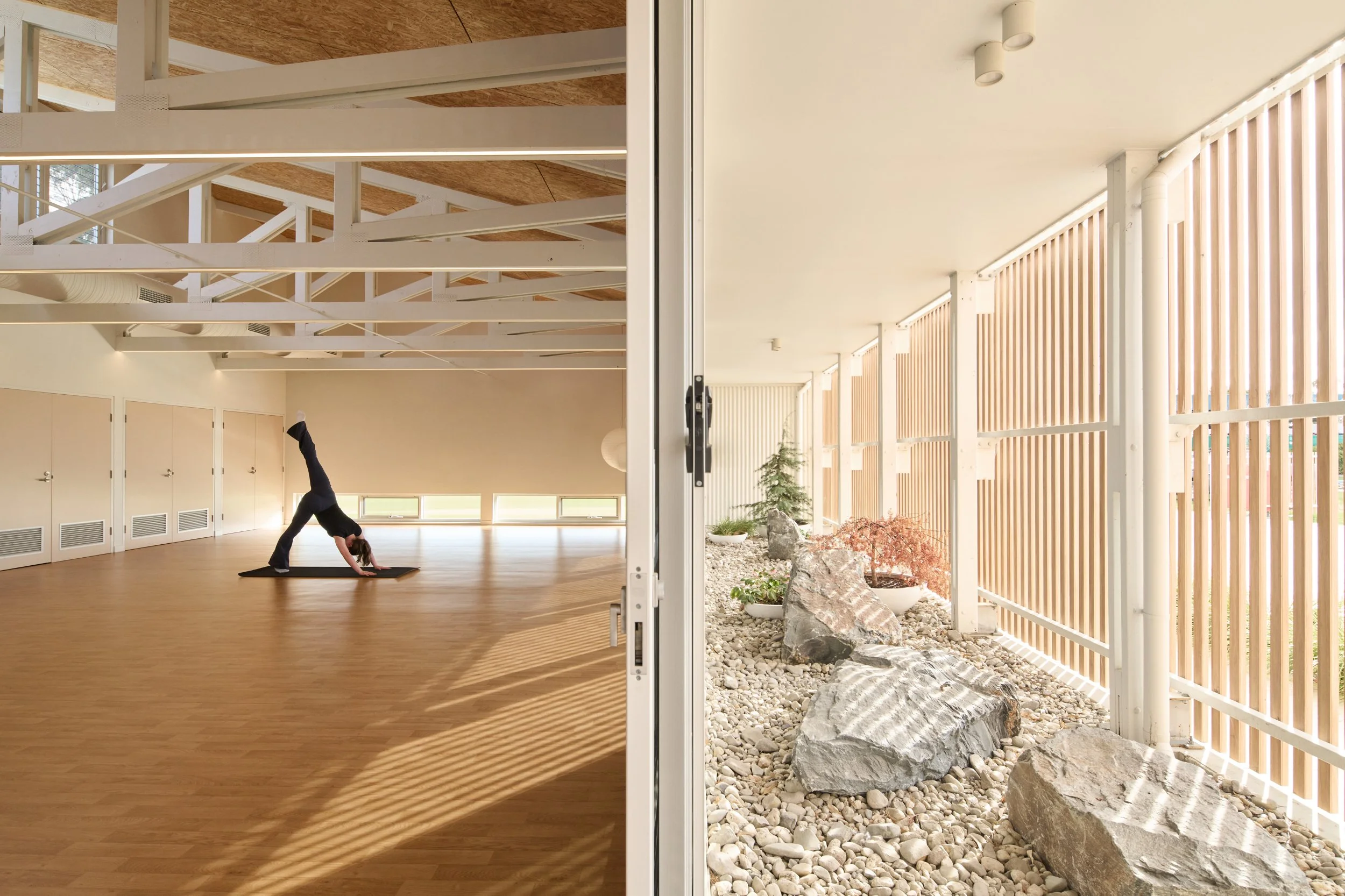 An open door leading to a room with a woman practicing yoga, indoor with wooden flooring and high ceilings, and a view outside of a courtyard with rocks and plants.