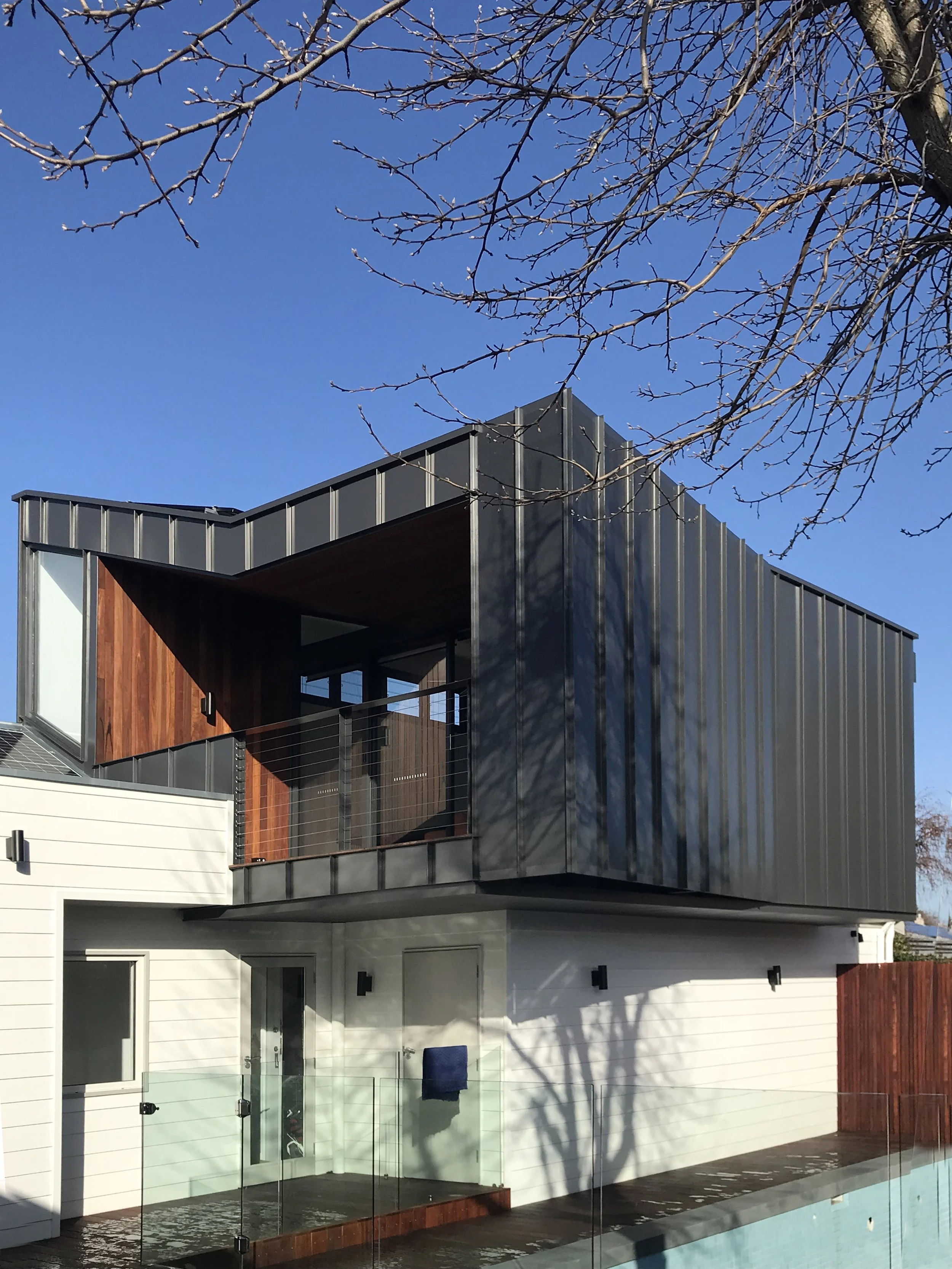Modern residential building with a balcony, glass railing, and outdoor pool, featuring a mix of wood and metal exterior walls, adjacent to a tree with bare branches, under a clear blue sky.