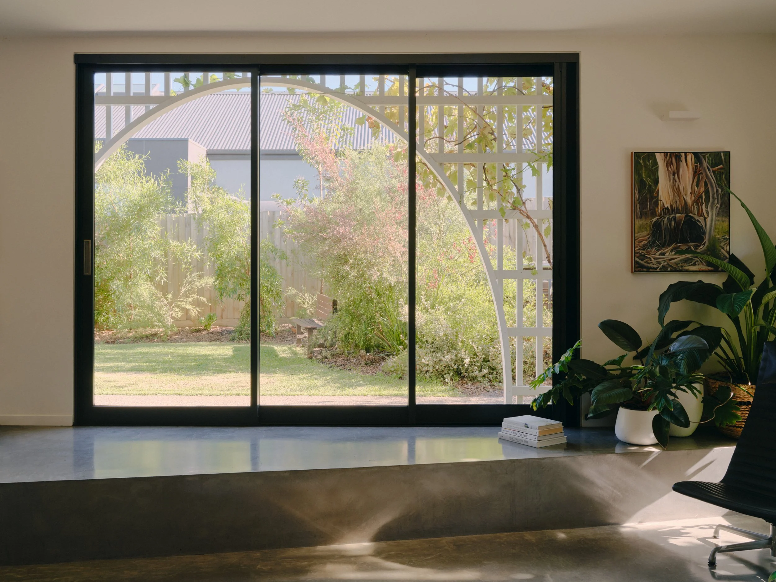 A modern living room with large glass sliding door opening to a lush garden with trees and a wooden bench outside. Inside, there is a tall green plant in a white pot, a chair, and a small stack of books on a white surface.