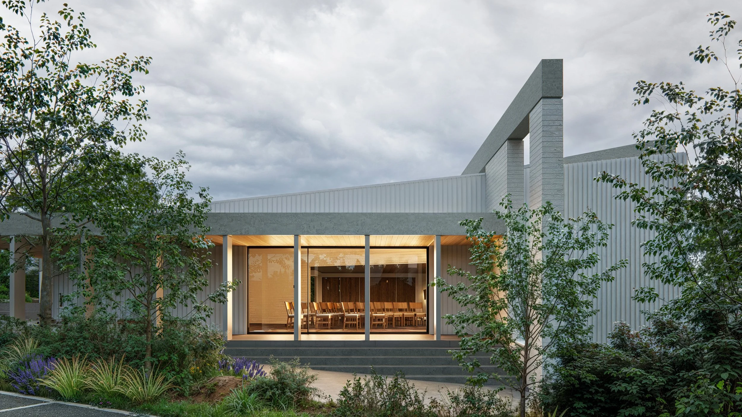 Modern building with large glass windows, surrounded by trees and greenery, under cloudy sky.