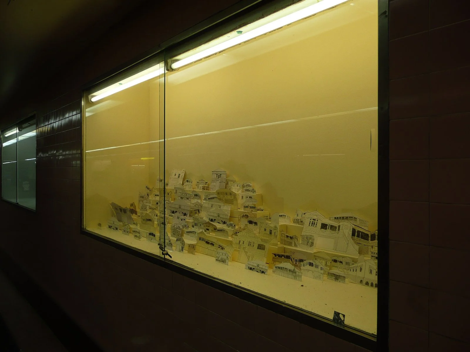 A display case with paper models of various buildings and houses, illuminated by bright lights, located in a corridor.