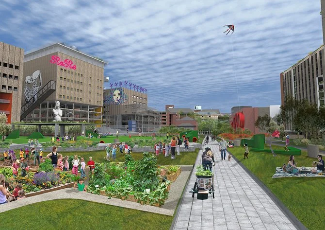 Urban park with people enjoying gardens, pathways, and outdoor activities in front of buildings with murals and signage, under a partly cloudy sky.