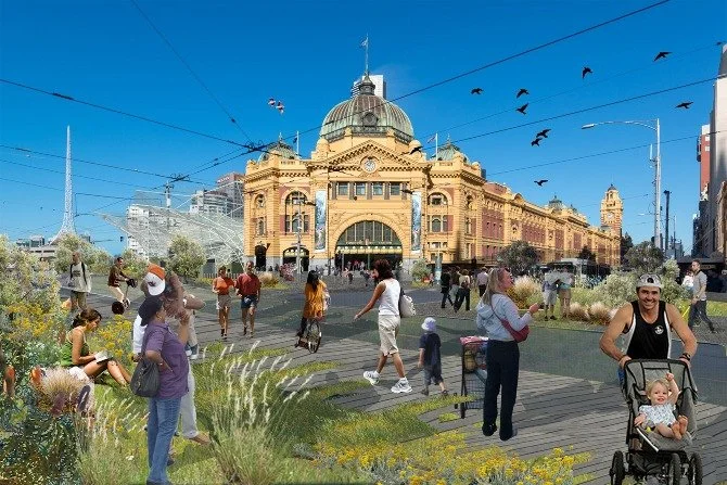 People walking and gathering in front of a historic train station with a large dome and arched entrance, under a clear blue sky, with birds flying overhead.