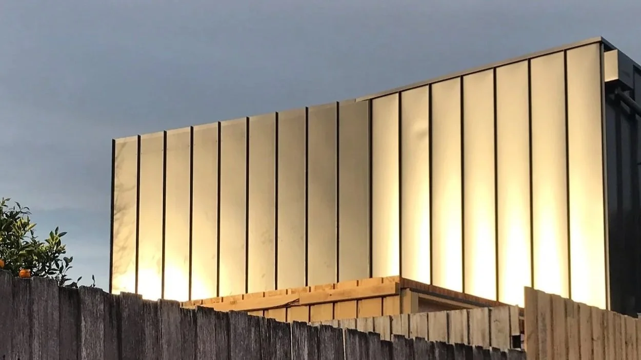 Exterior of a modern building illuminated at night, featuring tall vertical panels with backlighting and a wooden fence in the foreground.