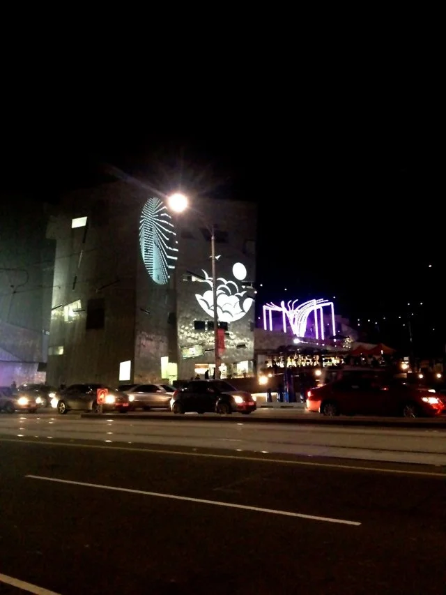 Nighttime view of a modern building with artistic light projections, including a large footprint and abstract shapes, with cars parked and driving on the street in the foreground.