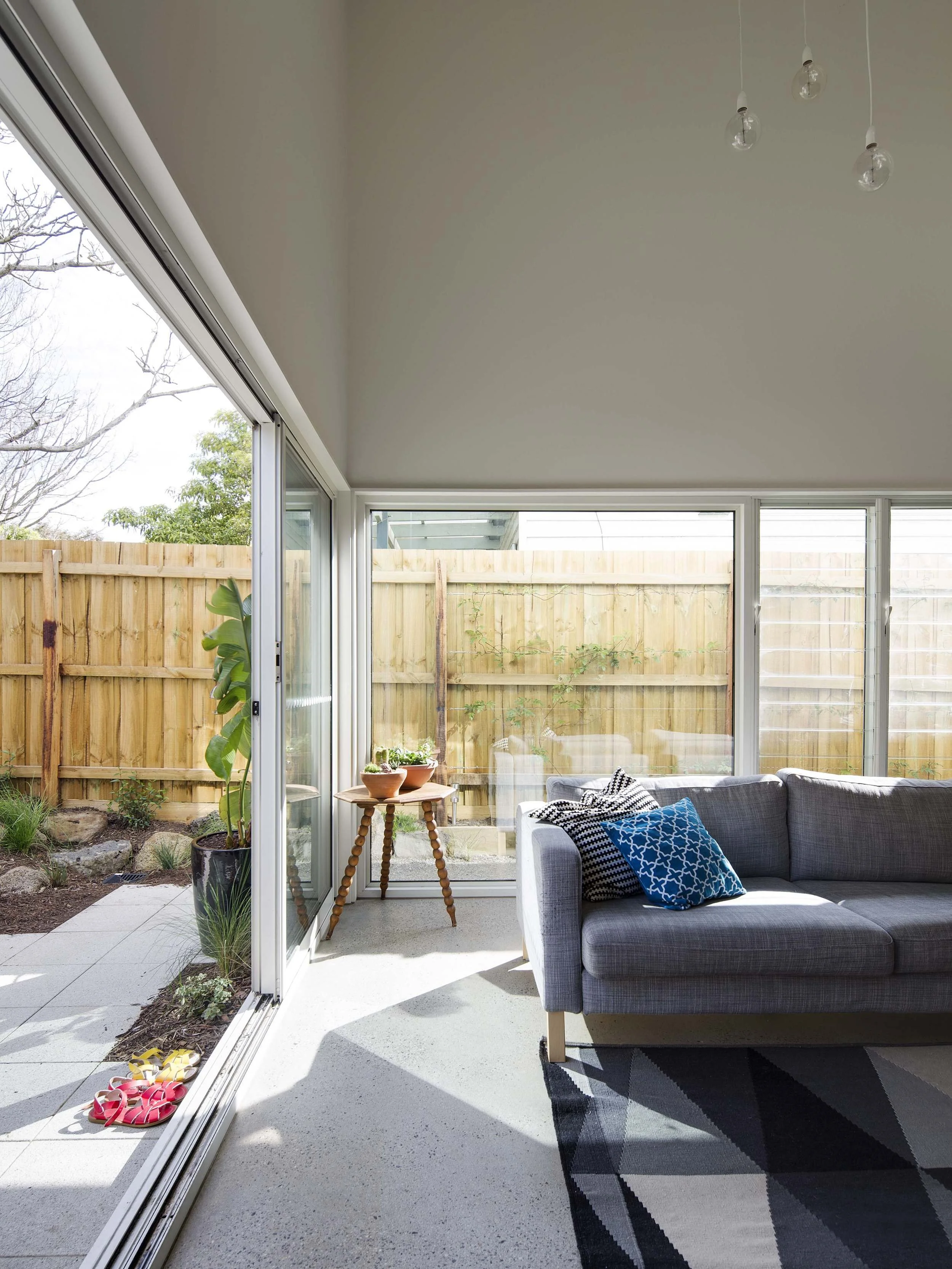 View of a bright sunroom with large sliding glass doors leading to a backyard with a wooden fence and some plants. Inside, there's a gray sofa with blue patterned pillows, a small wooden side table with plants, and colorful slippers on the floor. Han