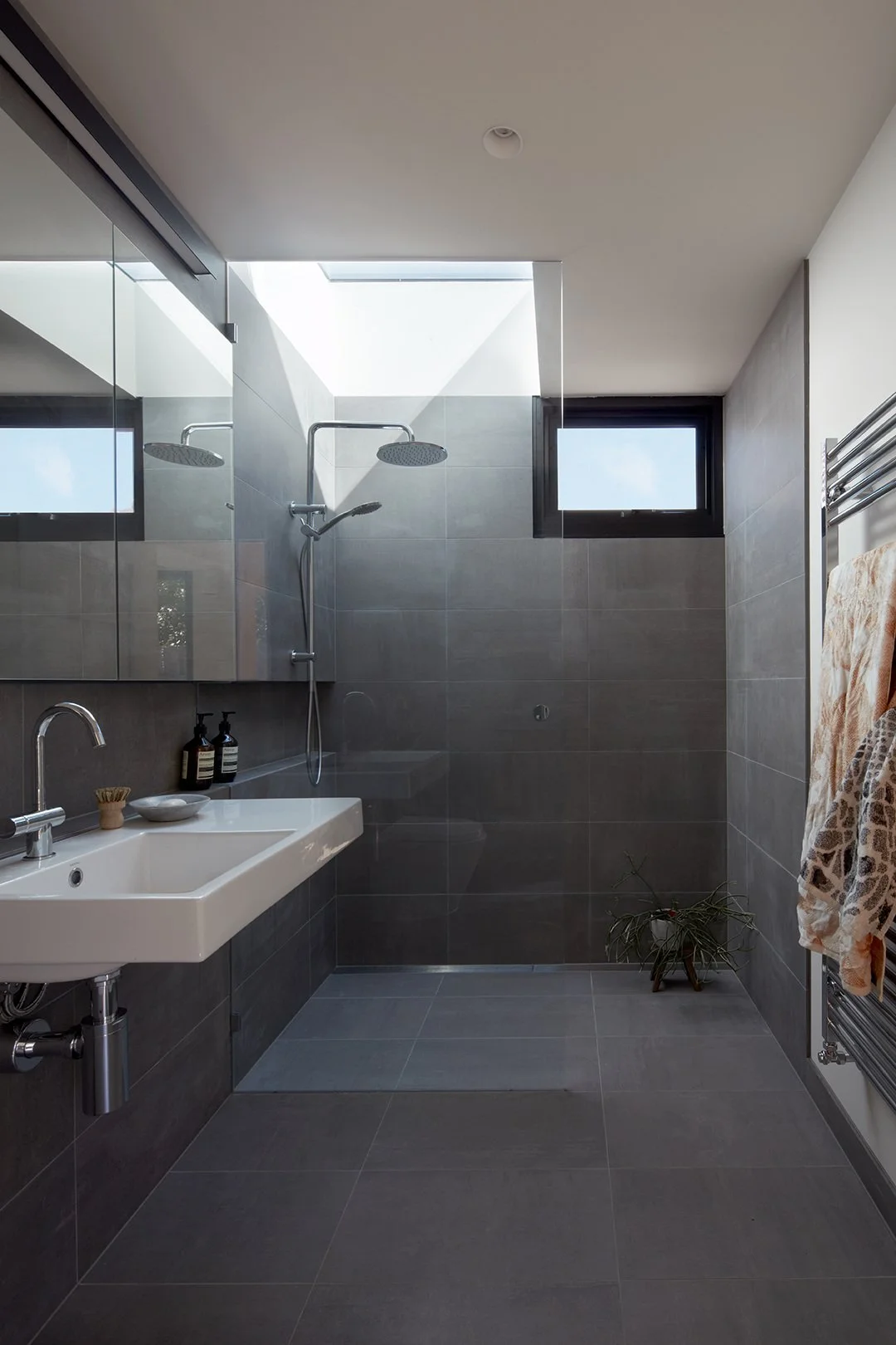 Modern bathroom with dark gray tiles, a white sink, black soap dispensers, a shower with a rainfall showerhead, a small window, a plant, and towels hanging on a heated towel rack.