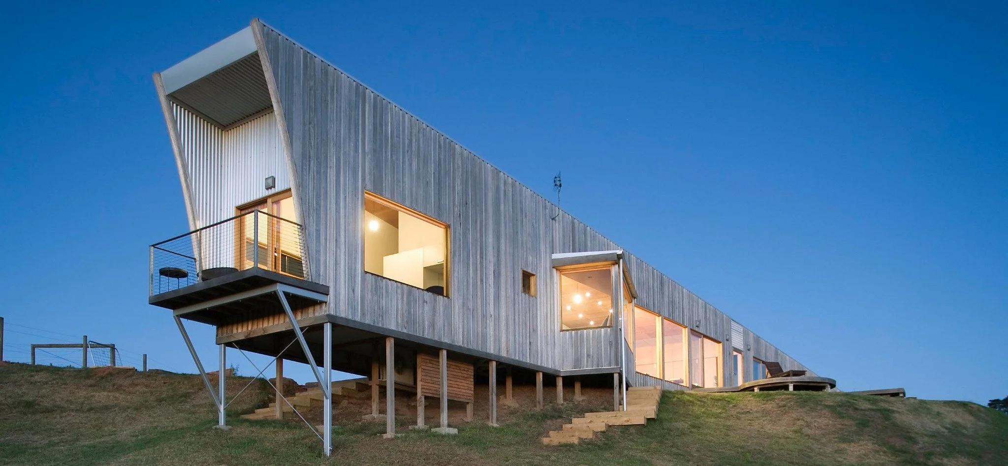 Modern wooden house on stilts with large windows and a balcony, built on a hillside under a clear evening sky.