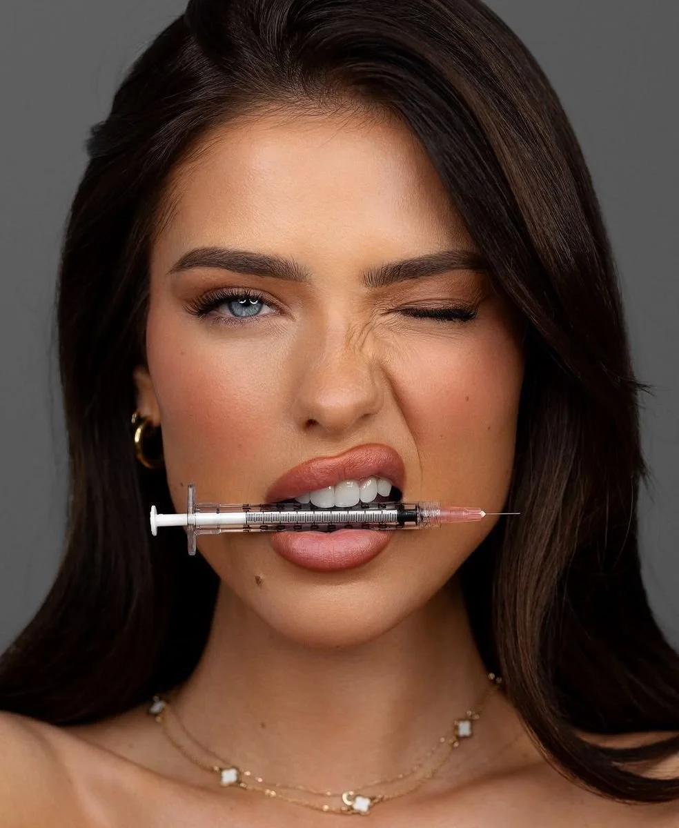 A woman with dark hair, blue eyes, and makeup holding a syringe in her mouth, winking with one eye, against a gray background.
