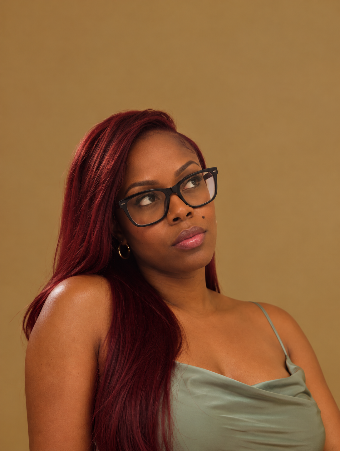 A woman with long red hair, wearing black glasses, gold hoop earrings, a mint green spaghetti strap top, looking upward.